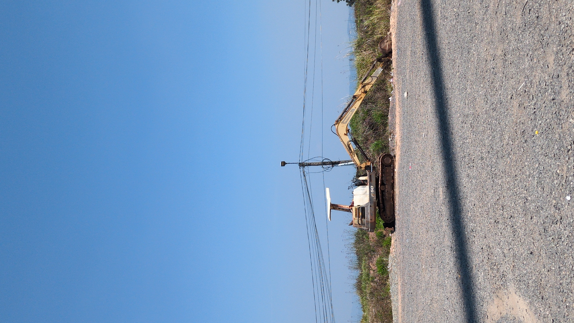 The image shows a yellow and white Yanmar F50 excavator parked on the side of a gravel road in what appears to be a rural or semi-rural area. The excavator is near a patch of grass and weeds. Above the excavator is a telephone pole with wires running along the road. The sky is clear and blue. This location is in Trường Thọ, Vietnam.