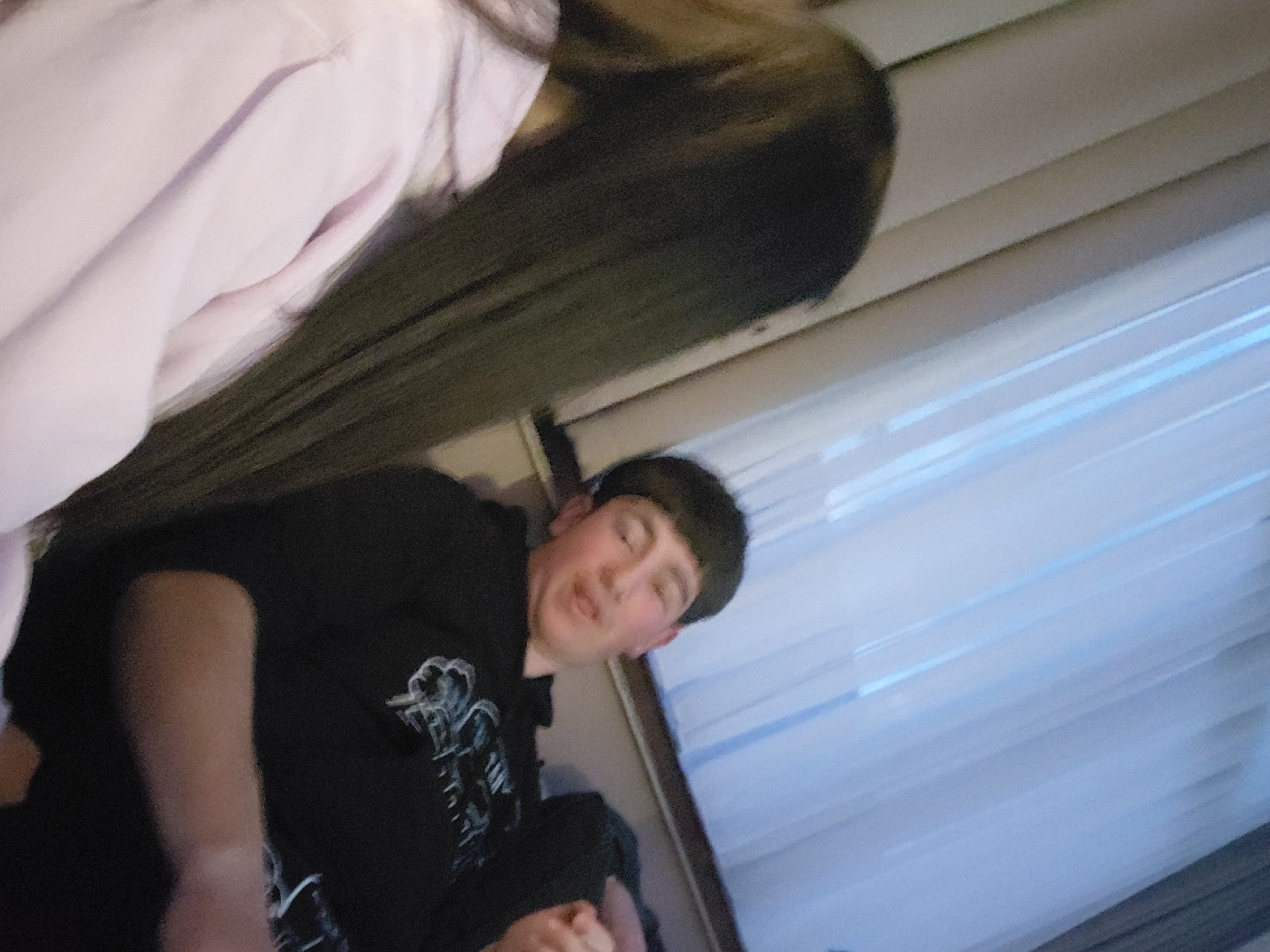 This candid, tilted photograph captures an indoor scene, likely in Kochan, Bulgaria, featuring two individuals. In the upper left quadrant, partially out of frame, is a person with long, dark hair, wearing a light pink or lavender long-sleeved garment. Their face is not visible.
To the right and lower in the frame is a young male with short, dark hair, wearing a black long-sleeved hoodie. The hoodie features a light-colored, abstract graphic design on its front. His eyes are closed or squinted, and his mouth is slightly open, suggesting a playful expression, possibly laughing or mid-speech. His hands are clasped or resting in front of him.
In the background to the right of the male subject, light-colored horizontal blinds are visible, possibly covering a window, with a faint blue light emanating from behind or through them. A light-toned wall or paneling is also visible. The lighting in the image appears somewhat dim, indicative of an indoor setting. The atmosphere conveyed is informal and personal. No discernible text is visible within the image.