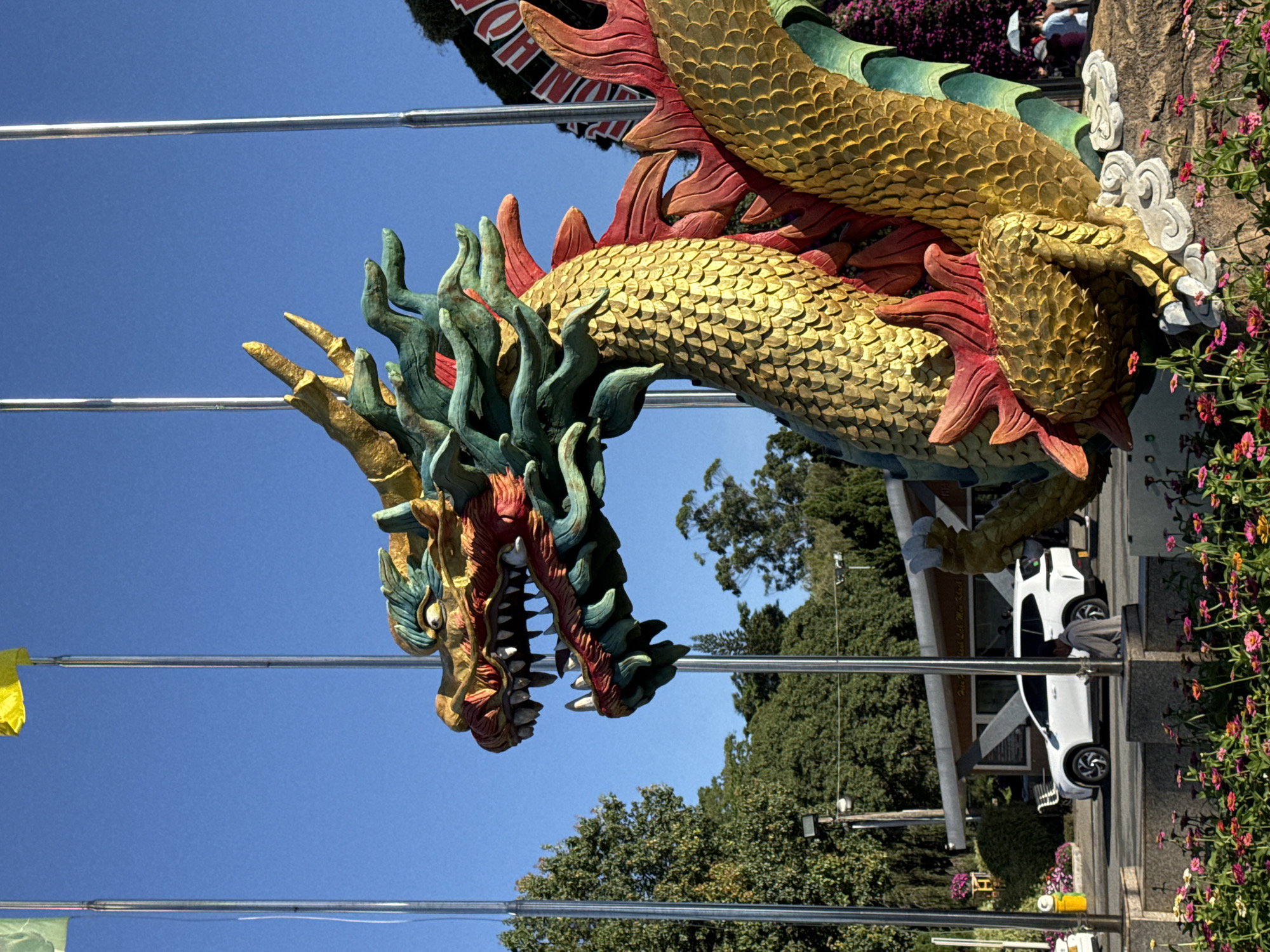 This is a medium-shot, eye-level, outdoor photograph featuring a large, colorful dragon statue in the foreground. The dragon statue is predominantly gold with accents of red and teal, and its scales are intricately detailed. The dragon's head is turned to the left, with its mouth open, revealing sharp teeth. Behind the dragon, several tall, silver flagpoles are visible against a clear blue sky. In the mid-ground, a white SUV is parked on a road, with a building and lush green trees in the background. The foreground is adorned with a vibrant display of pink and purple flowers. The scene is located in Phường Xuân Hương, Đà Lạt, Vietnam.