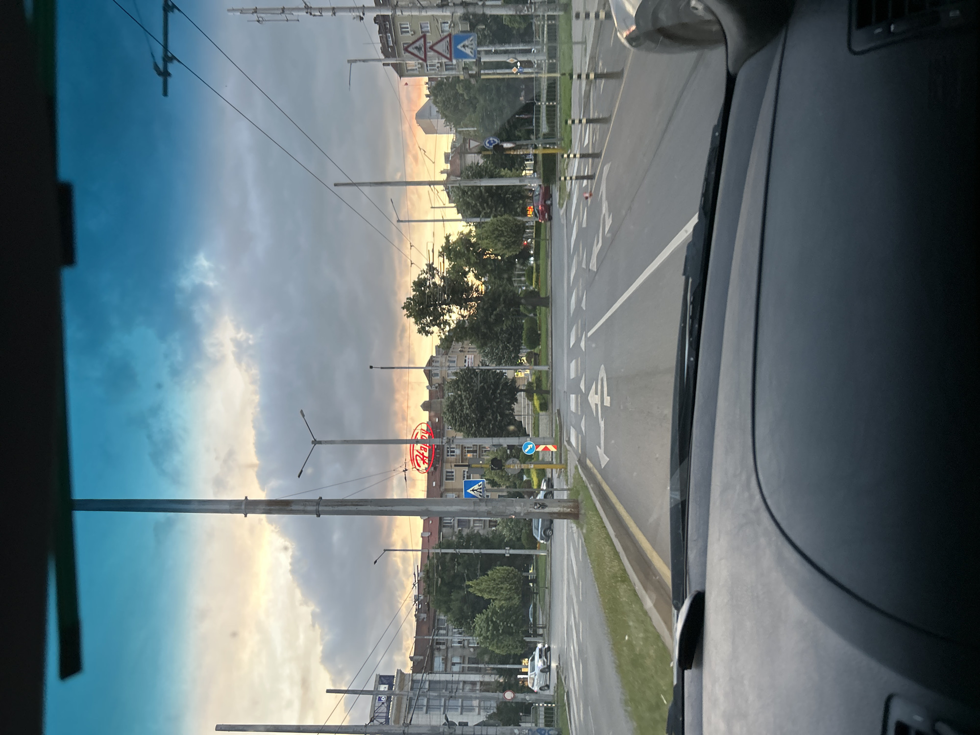 The image captures a view from the interior of a vehicle, looking out through the windshield at a city intersection during what appears to be dusk. The sky is a mix of blue and orange hues, with clouds scattered across it. In the foreground, the dashboard of the car is visible.
The intersection is busy with traffic. Several cars are present, including a white SUV in the left lane, a dark-colored SUV in the right lane, and a red car further down the road. Pedestrian crossings are marked on the asphalt, and traffic lights are visible. Streetlights and utility poles line the sides of the road, with electrical wires crisscrossing overhead.
Buildings of varying architectural styles, some appearing older and others more modern, form the backdrop of the scene. Trees and greenery are interspersed among the buildings and along the roadside, suggesting a well-established urban area. A neon sign, possibly for a business, is illuminated with red lights. Several traffic signs are also present, indicating directions and rules for drivers. The overall impression is that of an active urban environment transitioning from day to night.