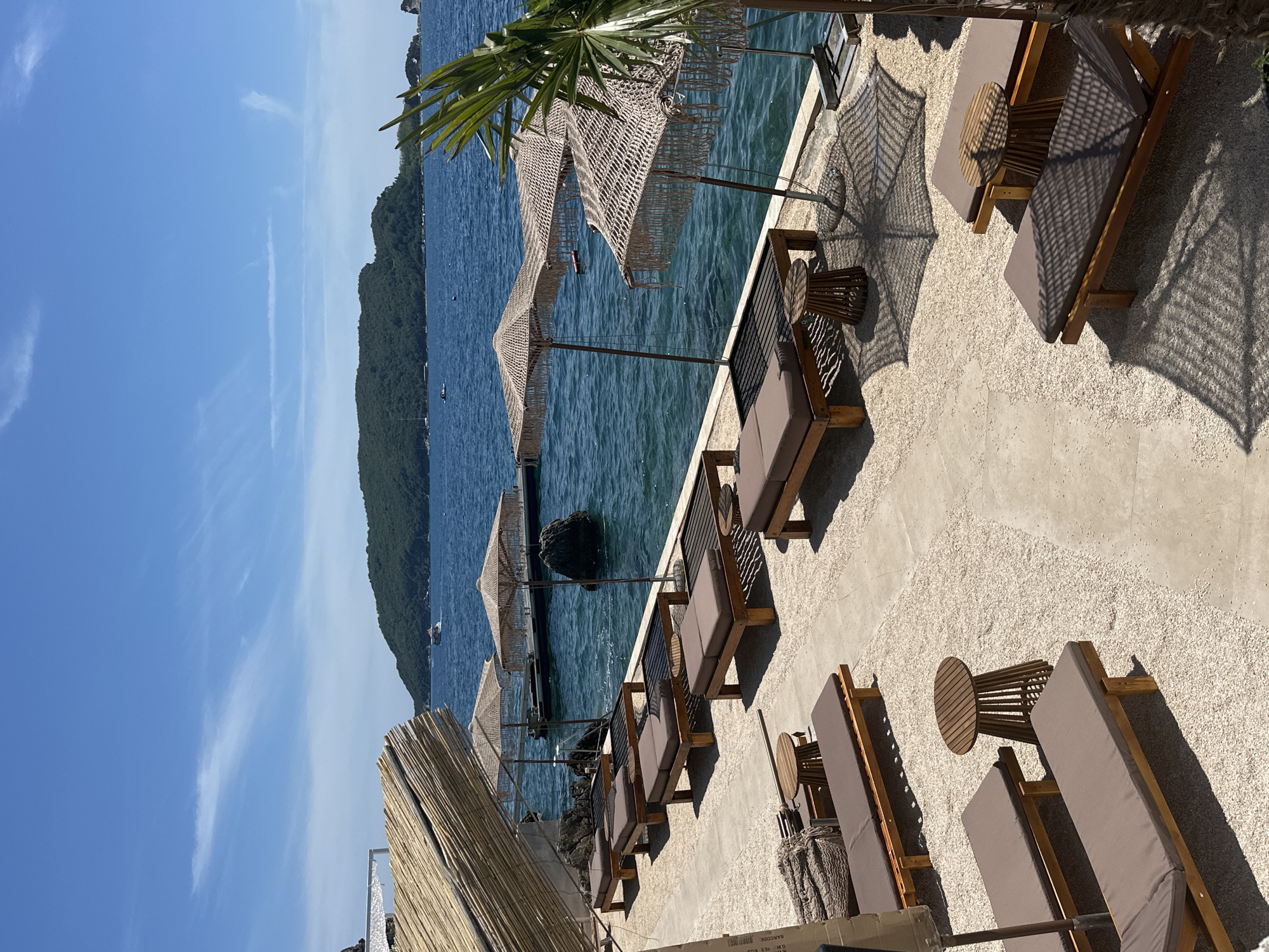 The image depicts a tranquil beachside scene in Budva, Montenegro. Numerous sun loungers with brown cushions are arranged on a light-colored gravel surface, some placed on concrete pathways. Several straw umbrellas with fringe details provide shade over some of the loungers. Small wooden tables are strategically placed between the loungers. In the background, the clear blue Adriatic Sea stretches out, with a lush green island visible in the distance. A few boats are dotted on the water. The architecture of the area includes a structure with a thatched roof. The overall atmosphere is serene and inviting, suggesting a resort or private beach club setting.