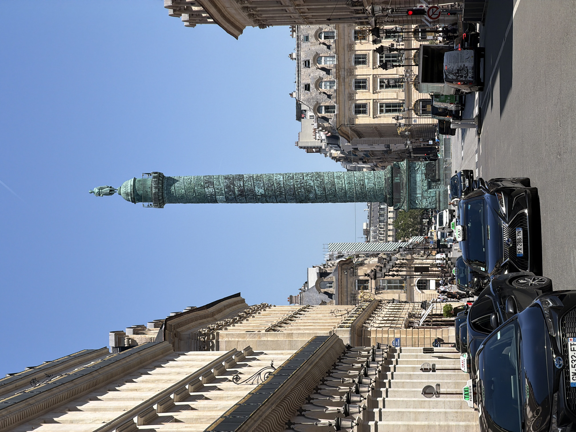 The image shows the Colonne de Juillet (July Column) in the Place de la Bastille in Paris, France. The fluted bronze column is topped with a gilded statue of the Génie de la Liberté (Spirit of Freedom). Buildings flank the column on either side. A street runs along the base of the column, with cars and taxis parked along the side. The sky is clear and blue. The image is taken from a low angle, looking up at the column.