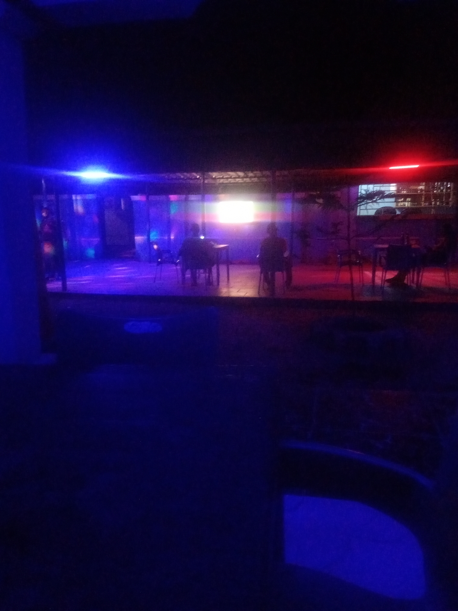 The image depicts an outdoor seating area at night, illuminated by various colored lights, suggesting a bar or entertainment venue. Two individuals are seated at separate tables, facing away from the camera. The environment appears to be somewhat rustic, with what looks like a corrugated iron roof structure overhead and simple tables and chairs. The overall ambiance is dimly lit and atmospheric, with prominent blue and red lighting. The location is identified as Sabon Gari, Nassarawa, Nigeria.