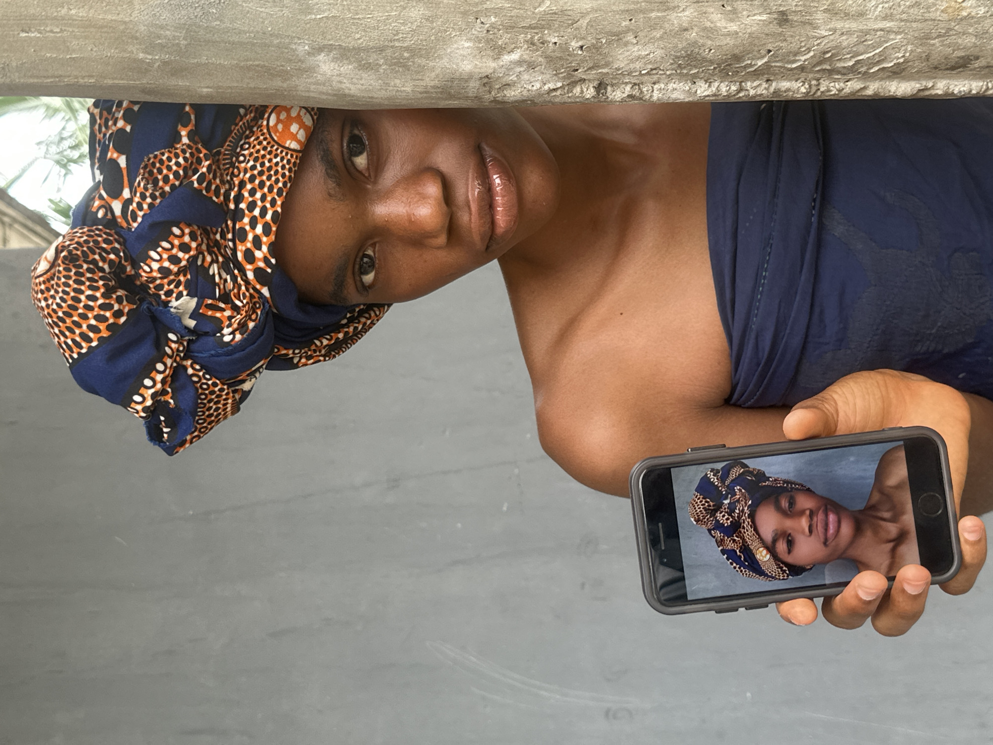 A young Black person with dark skin and eyes is holding up a smartphone in the foreground, displaying a selfie of themselves wearing a blue and orange patterned headwrap. The person's bare shoulders and upper chest are visible, and they are draped in a dark blue fabric. They are partially obscured by a rough, grey concrete column on the right side of the frame. Behind them, a grey, textured wall serves as the backdrop. The image appears to have been taken outdoors in Badagry, Nigeria, with natural light illuminating the scene.