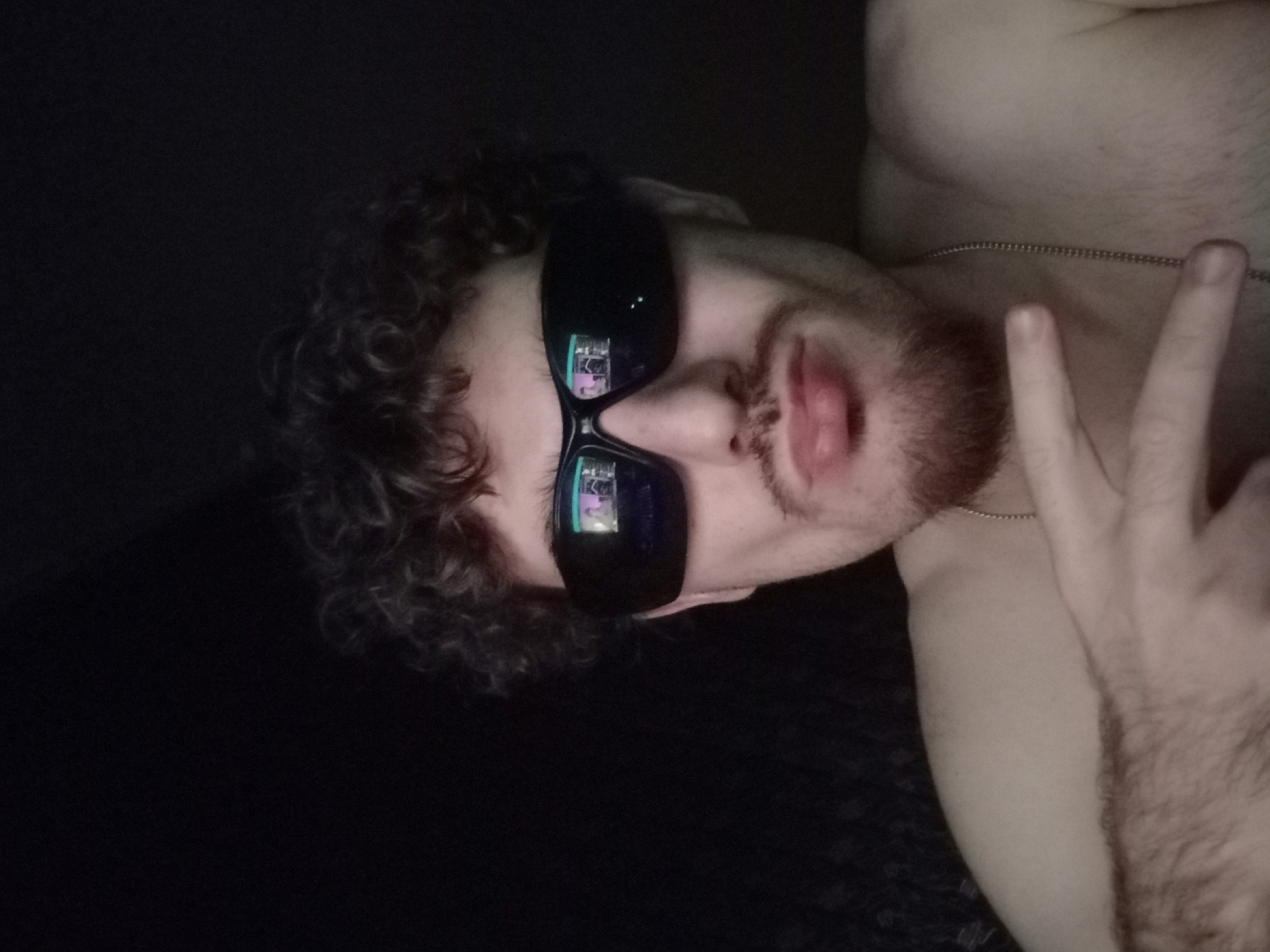 A close-up, waist-up photograph depicts a young man with curly, dark hair and a light beard, wearing dark sunglasses, against a dark background. The sunglasses reflect an image, possibly a screen or another person, showing figures and lights. The man is shirtless and wearing a silver chain. He is making a peace sign with his right hand, which is partially visible in the foreground. His left arm is also visible, covered in dark hair. The lighting is dim, with the primary light source appearing to come from the front, illuminating his face and upper body. There is no discernible location within the city of Parvomay, Bulgaria, visible in this image.