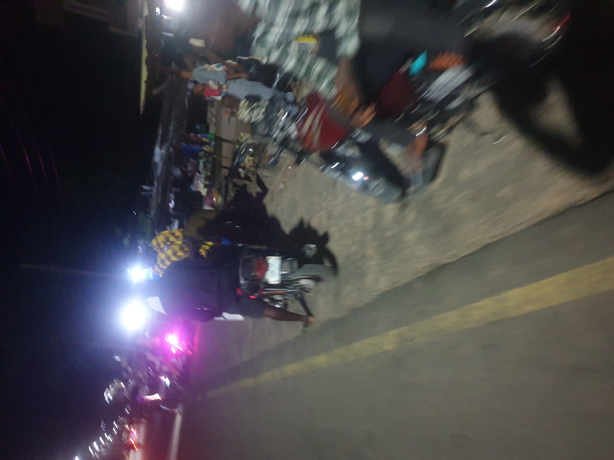The image depicts a night scene on a street in Zango-ndaji, Nigeria. Several motorcycles are parked along the side of the road, with some people sitting on them. One individual is standing near a motorcycle with a yellow and black checked pattern. The road has a yellow dividing line. Streetlights and other vehicle lights are visible in the distance.
