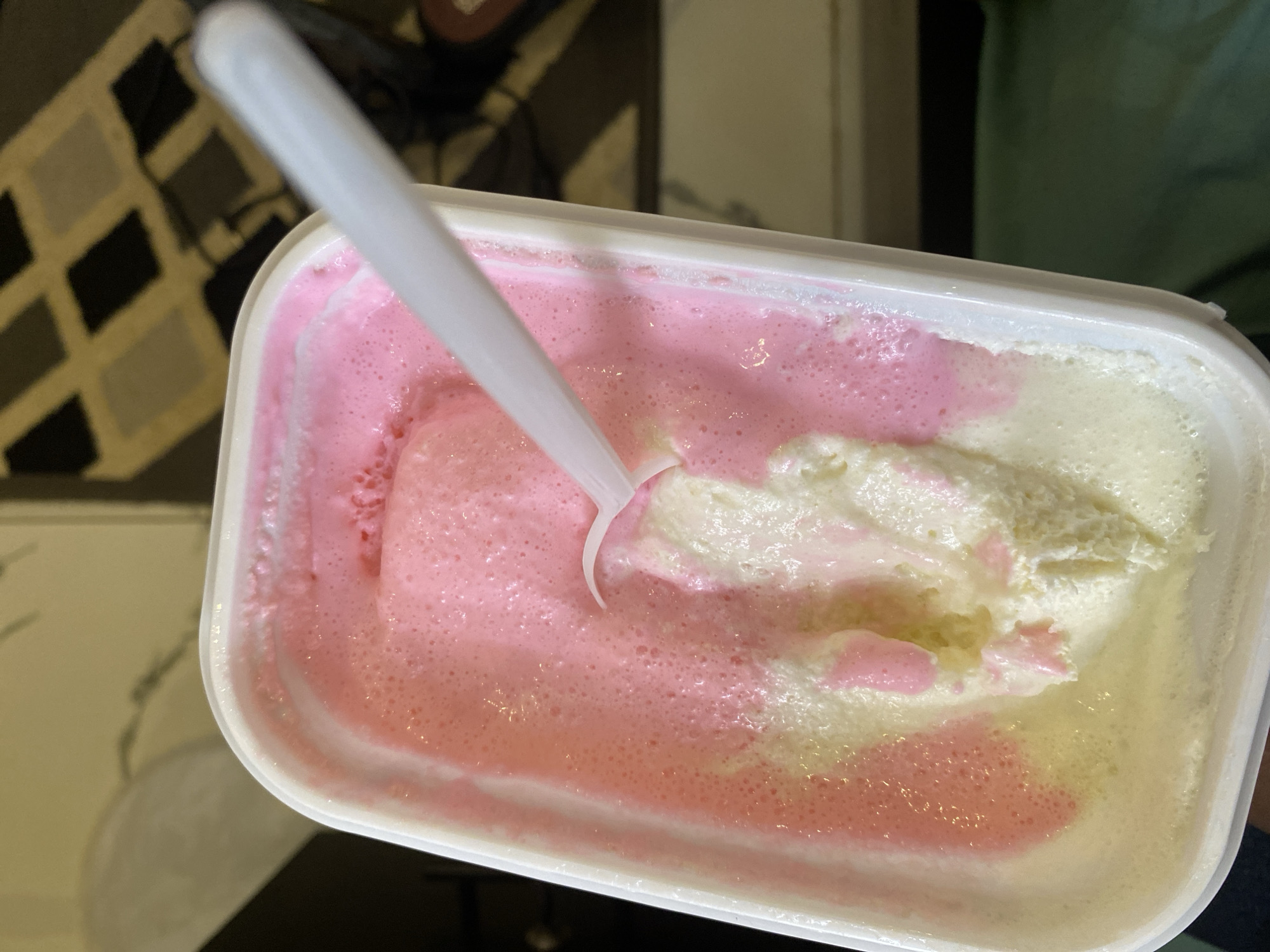 The image shows a close-up, overhead view of a white rectangular container filled with ice cream. The ice cream is a swirl of pink and white, with a white plastic spoon embedded in it, suggesting it has been eaten. The container is slightly tilted. The background is blurred, showing a patterned rug on the floor and a dark green cloth. There is no indication of the city of Dam Camp, Nigeria, in the image.