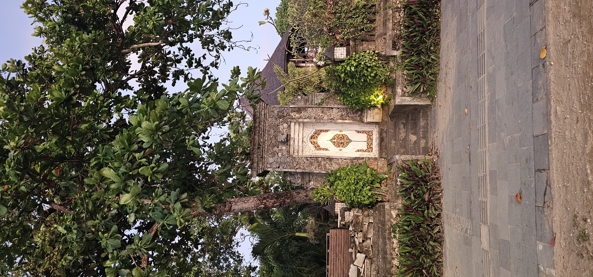 The image depicts a tropical scene, possibly a residential entrance, in Denpasar, Indonesia. An ornate, white double door with floral designs is set within a stone wall. Vegetation, including lush green foliage and tropical plants, surrounds the doorway. A pathway paved with gray stone leads to the entrance. A tree with broad, green leaves dominates the left side of the frame. The lighting suggests it is either early morning or late afternoon.