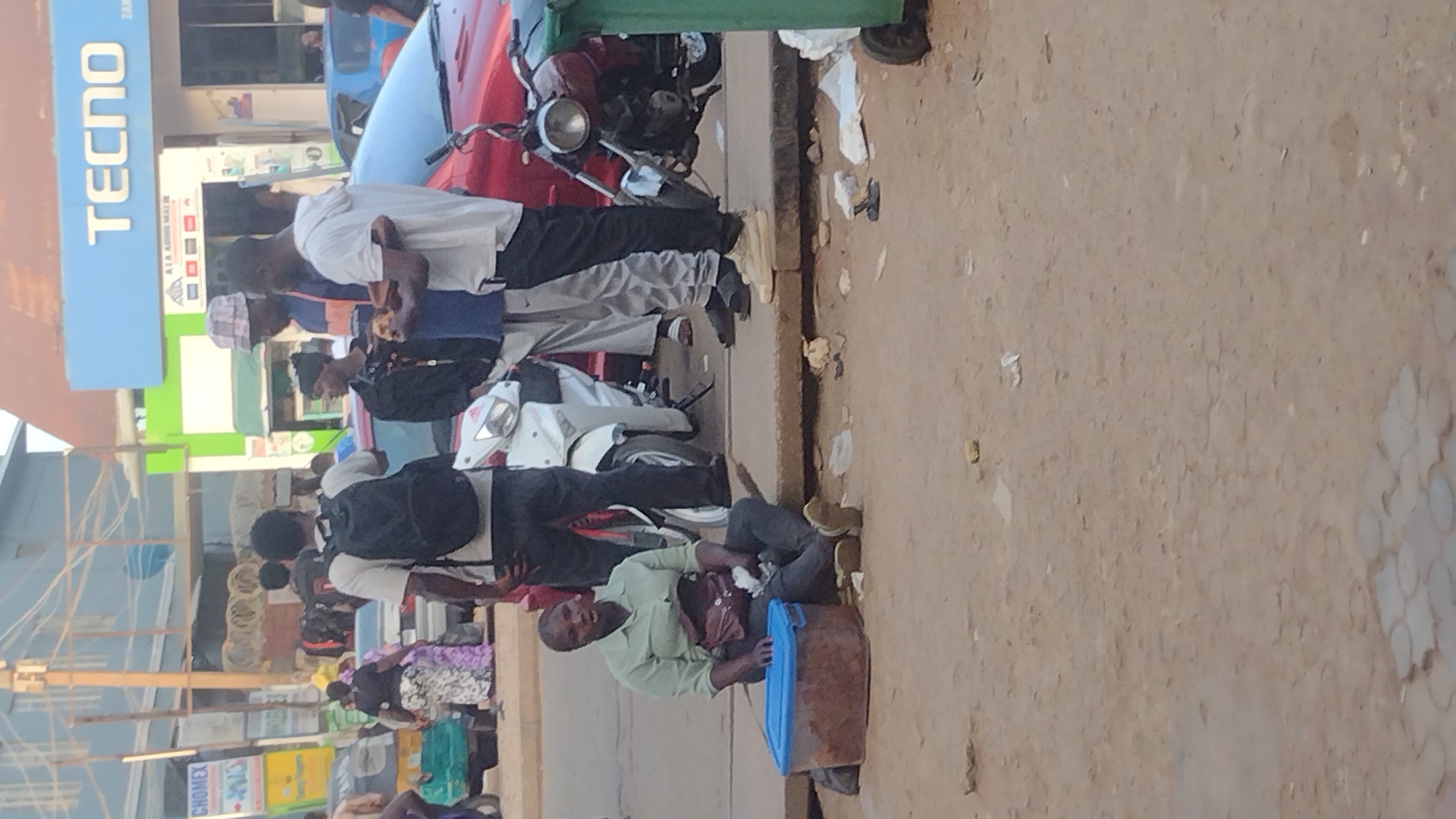 The image shows a street scene in Jos, Nigeria. Several people are standing and sitting near a curb. One man is seated on a container, apparently selling something, as he has a bag in his lap. Several others are standing nearby, some eating. There are parked motorbikes and a car adjacent to the curb. Signage for 'TECNO' and 'CHOMEX' are visible on buildings behind the people. The ground is bare and appears to be slightly uneven.