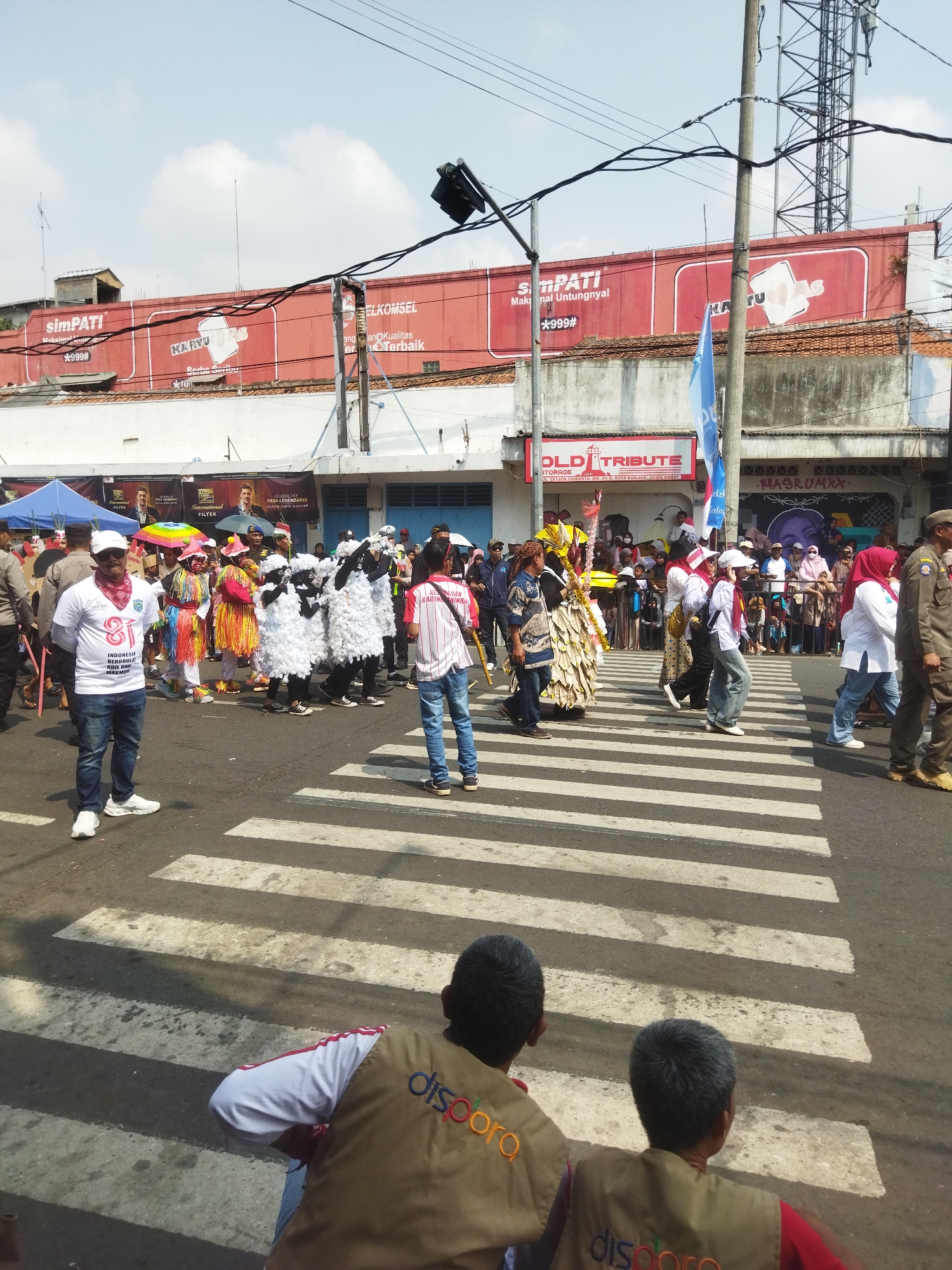This image depicts a street scene in the city of Banjar, Indonesia, likely during a parade or festival. A group of people dressed in elaborate costumes, including individuals in white sheep-like outfits and others in colorful, fringed attire, are crossing a pedestrian zebra crossing. One person in a white t-shirt and blue jeans stands prominently on the left side of the crossing, facing the viewer. Several other individuals, some in uniforms, are also visible in the background, along with spectators lining the street. Overhead, power lines and a communication tower are present, and buildings with large red billboards, one advertising "simPATI," are visible in the background. The sky is clear and blue.