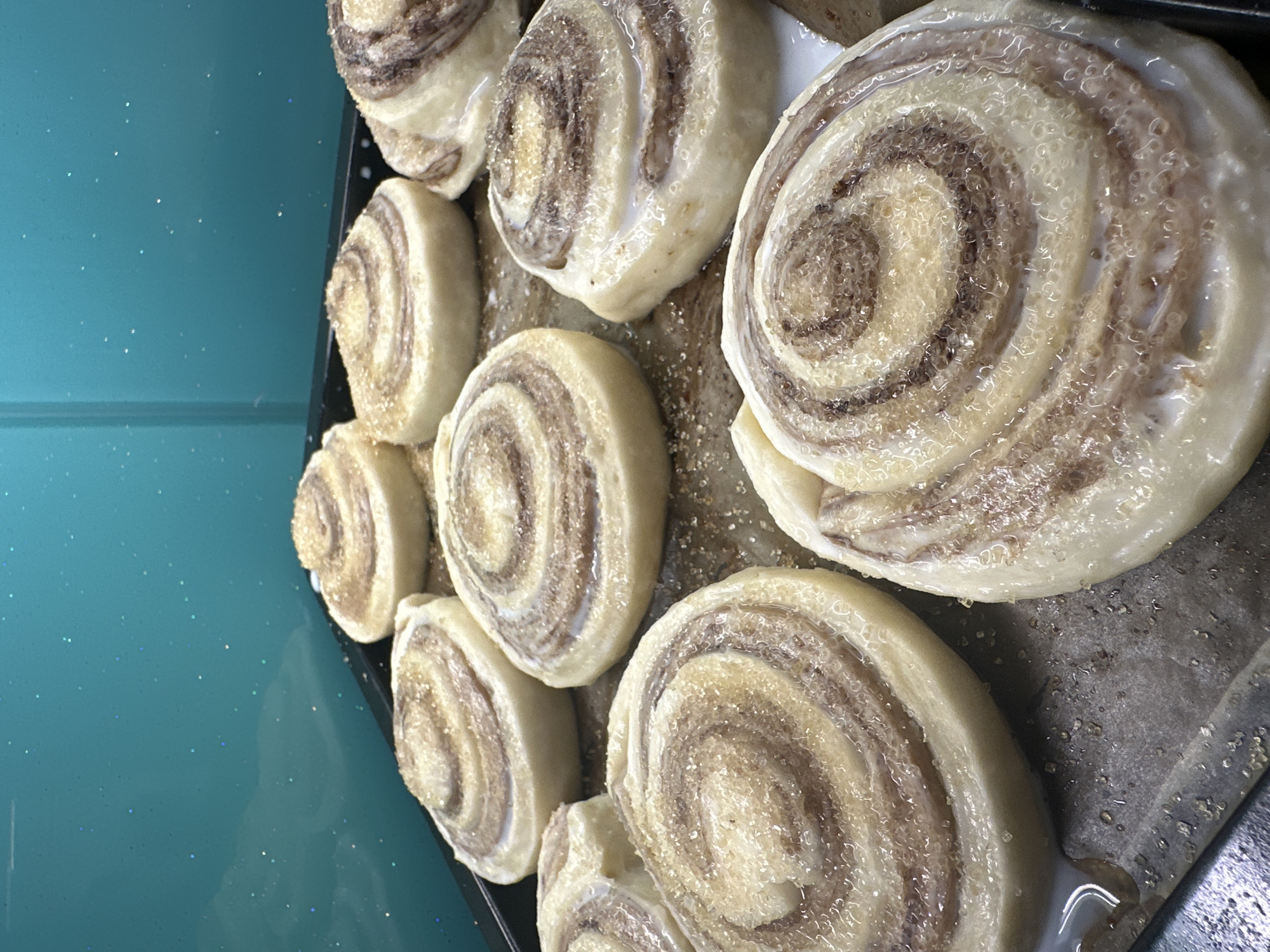 A black baking pan contains a cluster of cinnamon rolls, some larger than others, with visible swirls of brown and white filling. The rolls are coated with a sugary glaze. The pan is placed on a reflective, turquoise surface. The image's angle suggests it may have been taken in a kitchen or food preparation area, but no specific location in Bac Nha Trang Ward, Vietnam can be determined from the visual information alone. There are no people visible in the image.