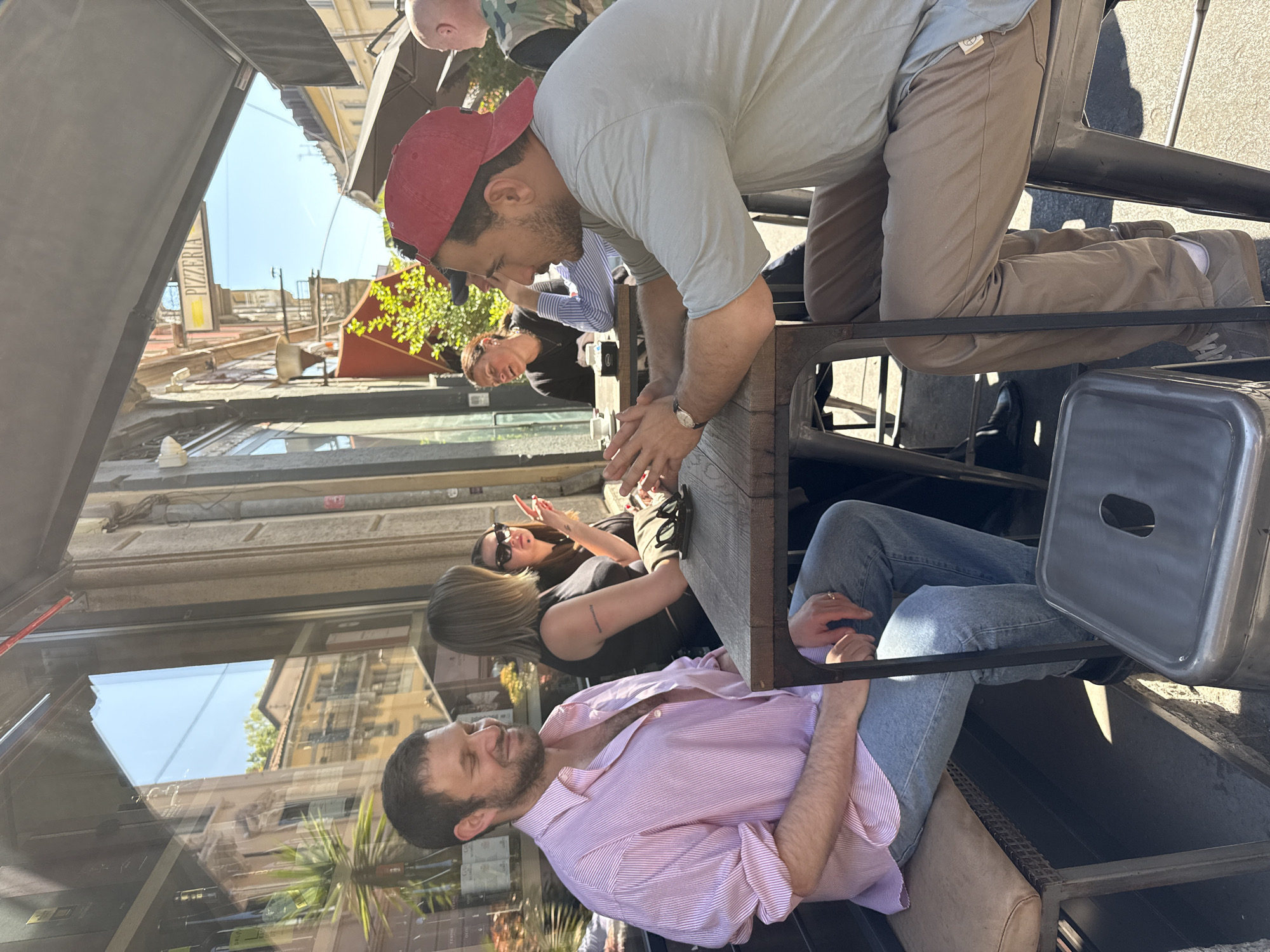 The image captures an outdoor scene in Milan, Italy, likely near a restaurant or café. Several people are seated at a wooden table, including a man in a pink striped shirt, another man wearing a red cap, and a woman with sunglasses. The architectural details visible through the window reflect Milan's urban landscape.