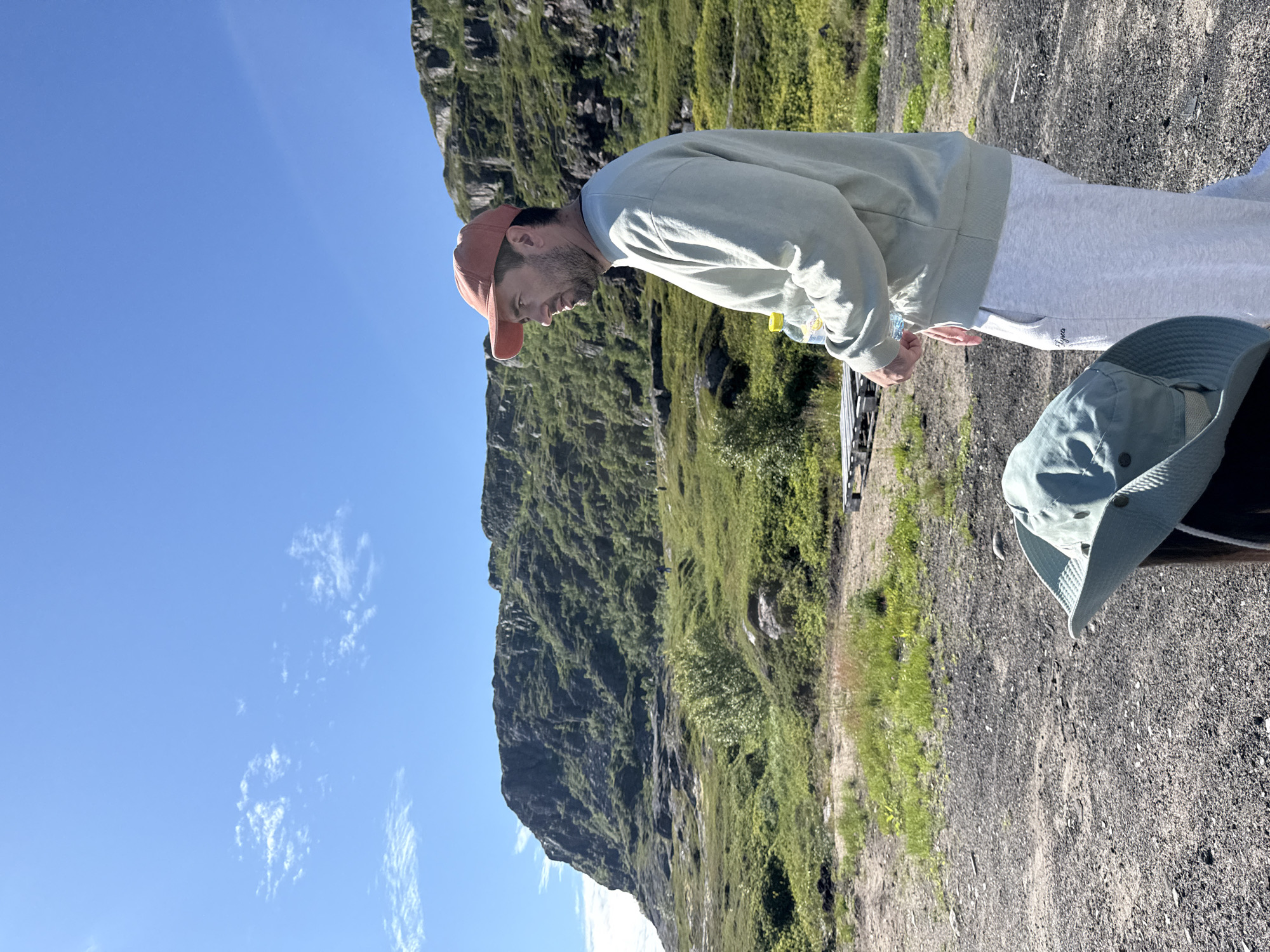 A man in a light green sweatshirt, light grey sweatpants, and an orange baseball cap stands in the foreground, facing right and looking away from the camera. He holds a clear plastic water bottle with a yellow cap in his right hand. In the lower left corner, a light green bucket hat rests on a dark cylindrical object. The background features a rocky, barren terrain with sparse green vegetation leading up to a large, steep, vegetated cliff face. The sky above is clear and blue with a few scattered white clouds. The scene appears to be outdoors in a natural landscape, possibly in or near Teriberka, Russia, given the rugged terrain and coastal cliff formations.