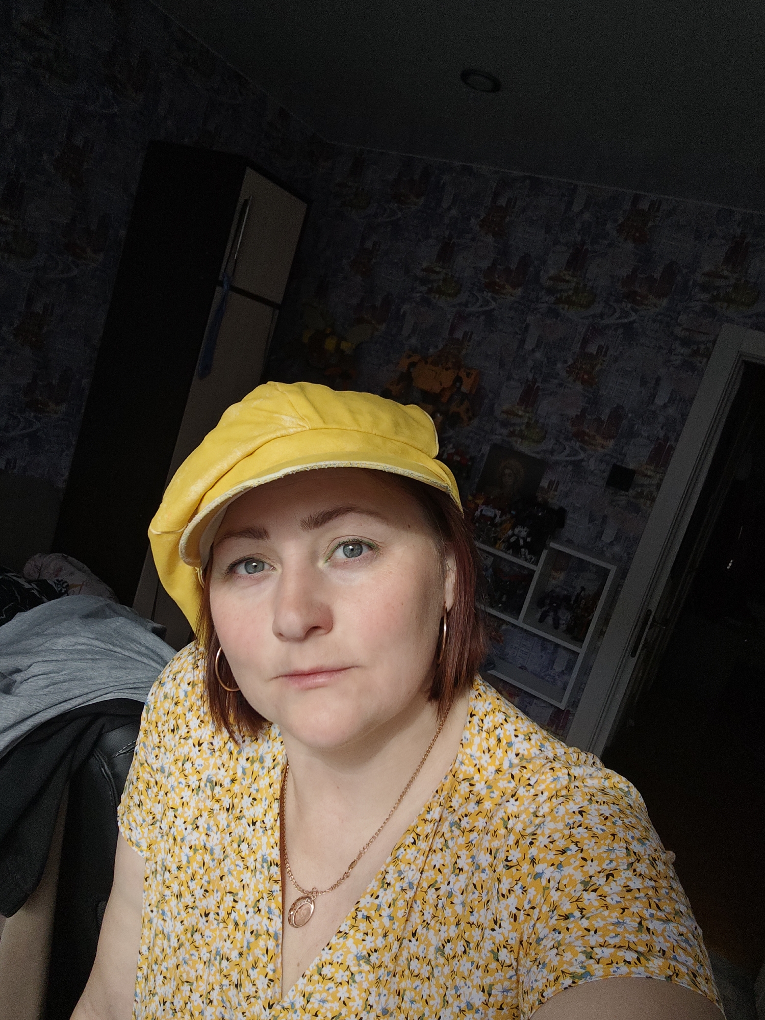 A woman is shown in a medium close-up shot. She is wearing a yellow newsboy-style cap and a yellow floral print dress with a V-neck. She has short auburn hair, light skin, and is wearing gold hoop earrings and a gold necklace with a pendant. Her gaze is directed toward the camera with a neutral expression. Behind her is a room with dark, patterned wallpaper. A tall, dark cabinet or refrigerator is visible to the left. A white shelving unit with what appear to be figurines or collectibles sits against the wall behind her right shoulder. A doorway is visible in the background to the right. The location is Markovskii, Russia.