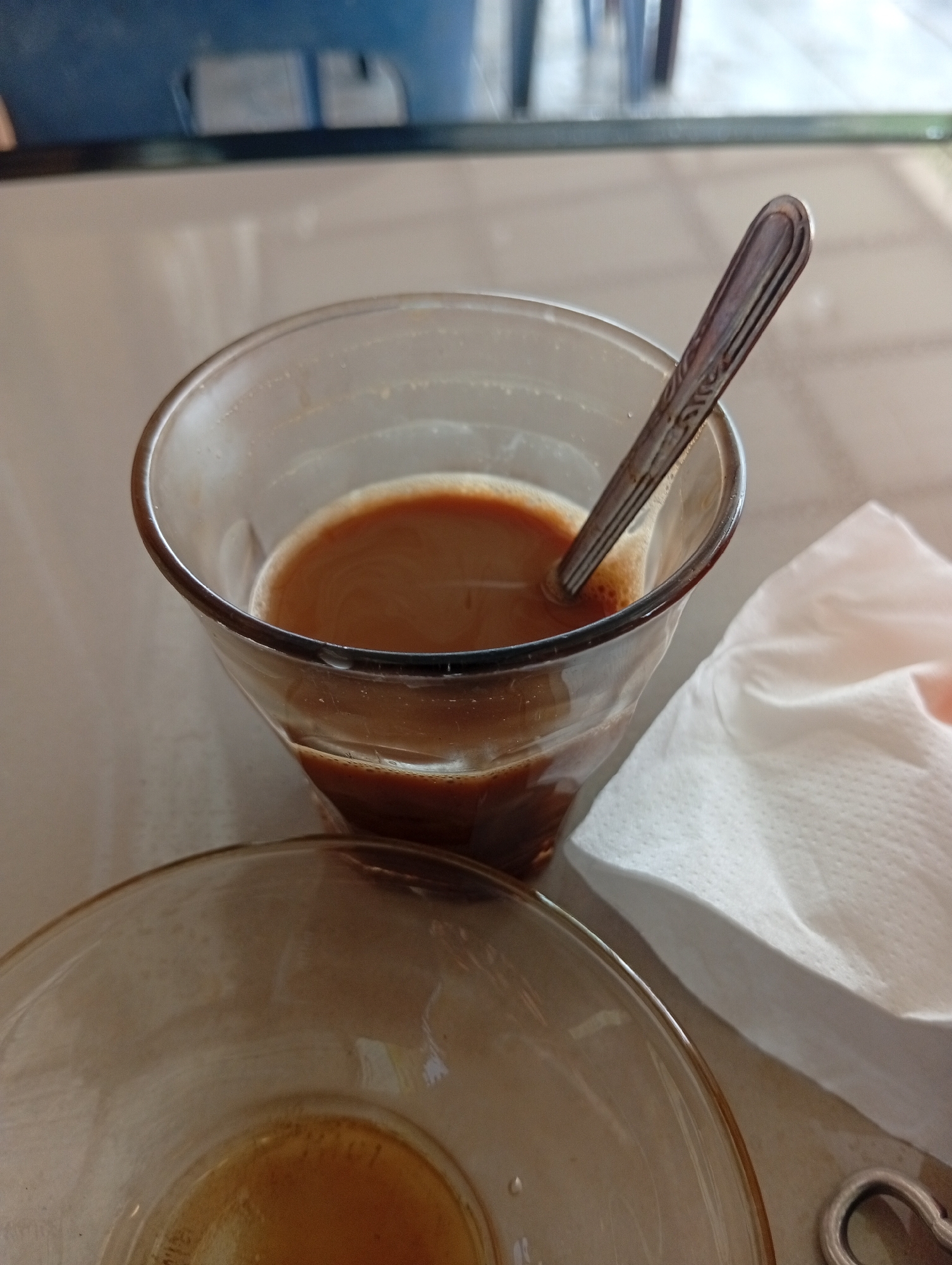 A close-up, high-angle shot captures a glass of coffee with a spoon inside, placed on a table. To the right of the glass, a white paper napkin is folded. A second, empty glass is visible in the foreground, partially obscuring a portion of the table surface. The background is blurred, but appears to show furniture and possibly a window. The scene is likely set in a cafe or restaurant in Lhokseumawe, Indonesia.