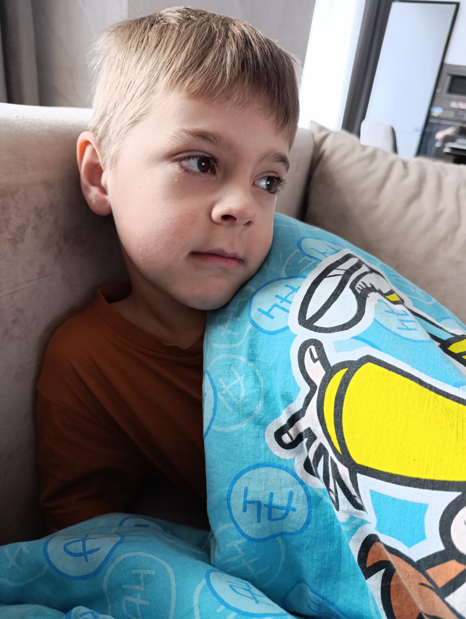 A young boy is pictured indoors, seated comfortably and partially covered by a light blue blanket. He has light brown hair, fair skin, and dark eyes, and is wearing a simple brown t-shirt. His expression appears pensive or reflective, with his gaze directed to the right of the frame, and a slight wetness is visible at the corner of his left eye, suggesting tiredness or a quiet emotion.
He is leaning against a light-colored, upholstered sofa or armchair, which occupies the background. The setting is clearly a domestic interior, possibly a living room or bedroom. The background is brightly lit and out of focus, revealing what seems to be a large window or light source, along with indistinct elements of a room, possibly furniture or appliances. The bright lighting suggests it is daytime.
The light blue blanket covering the boy features a cartoon-style design, including a prominent yellow, black, and white illustration of what appears to be a character or part of a vehicle. Additionally, the blanket is patterned with light blue outlines of abstract shapes and speech or thought bubbles. Within some of these bubbles, the Cyrillic letters "ЧЧ" are clearly visible. These letters, pronounced 'Ch', are notable as they are the first letters of "Chaikovsky," the provided location context. The overall scene conveys a sense of quiet repose or introspection.