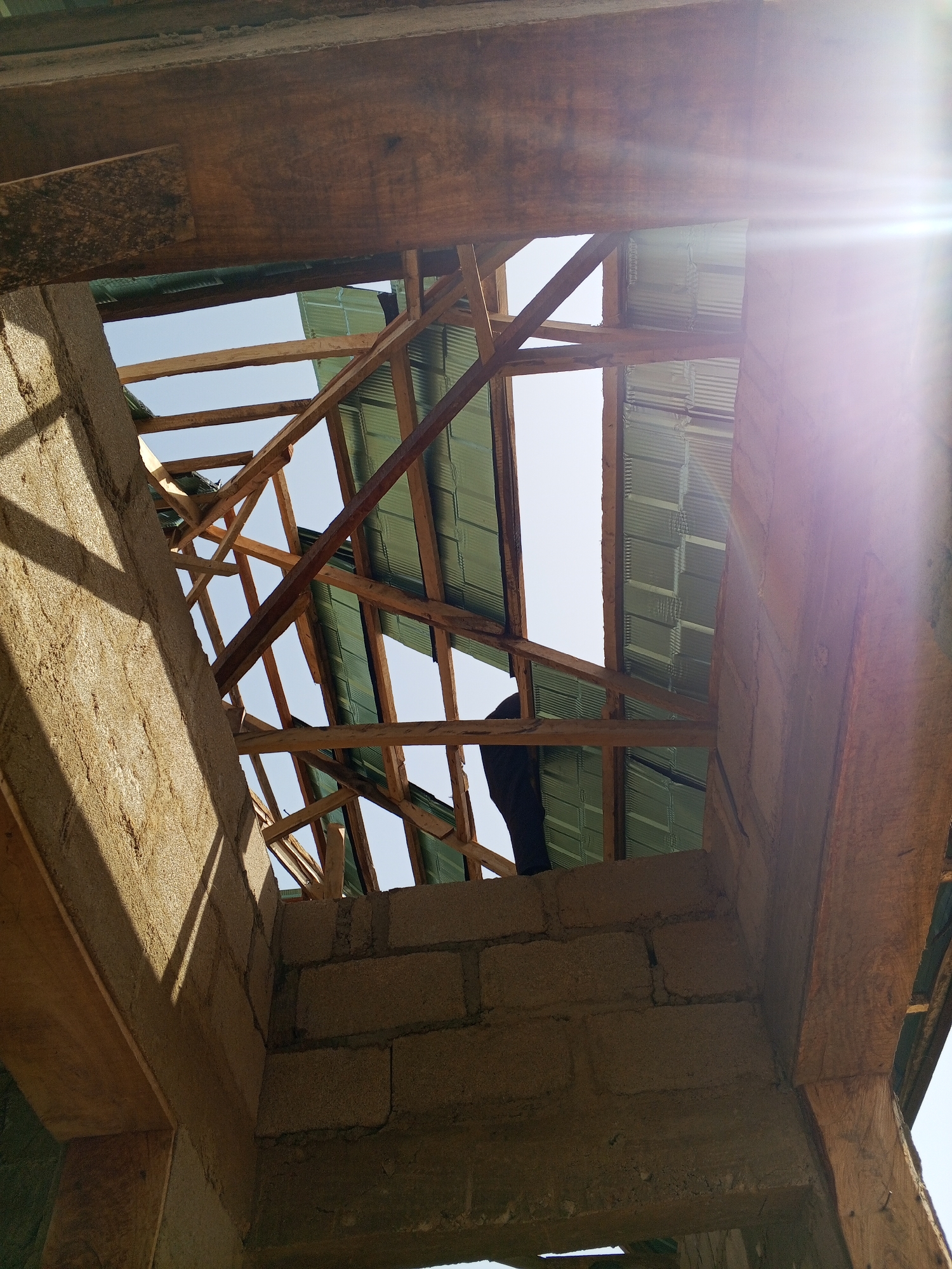 This image shows an unfinished building under construction. The walls are made of concrete blocks, and the roof structure is partially built with wooden beams and corrugated metal sheets. The sky is visible through the openings in the roof. The location is listed as the city of Unknown, Unknown. There is a person standing on the roof.