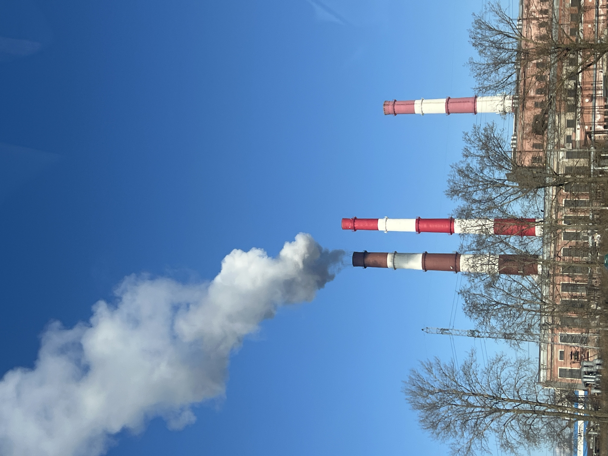 The image shows three smokestacks of a power plant in Saint Petersburg, Russia, against a clear blue sky. From one of the smokestacks, white smoke is billowing out. The smokestacks are painted with alternating red and white stripes. Bare trees are visible in front of the power plant building. A crane is partially visible behind the trees. The building is brick with rows of windows.