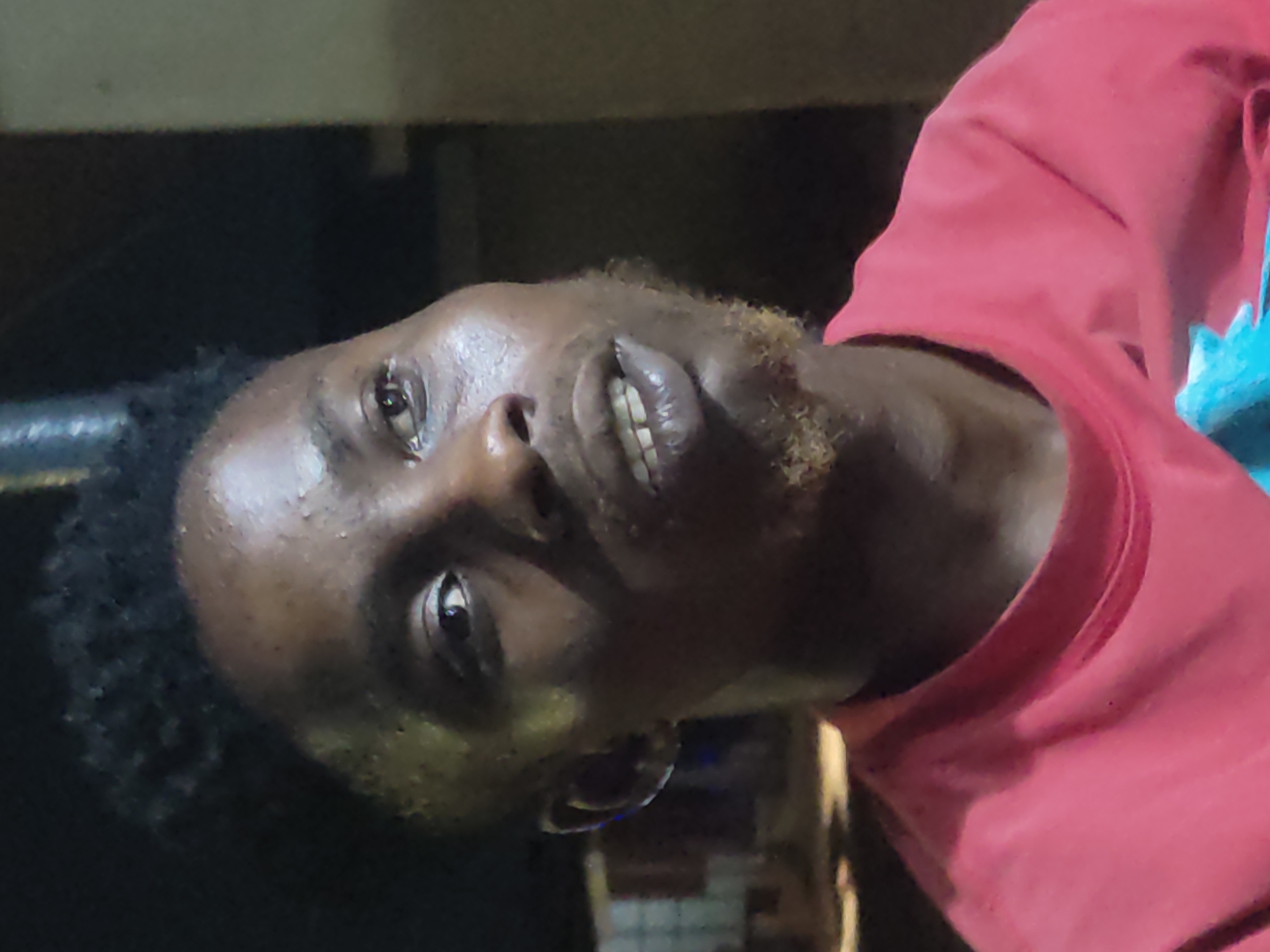 A close-up, slightly underexposed, portrait shot features a dark-skinned individual wearing a red long-sleeved shirt. The person has short, dark curly hair and some light facial hair on their cheeks. The background appears to be dark and indistinct. The location is alleged to be Ikorodu, Nigeria, based on the prompt.