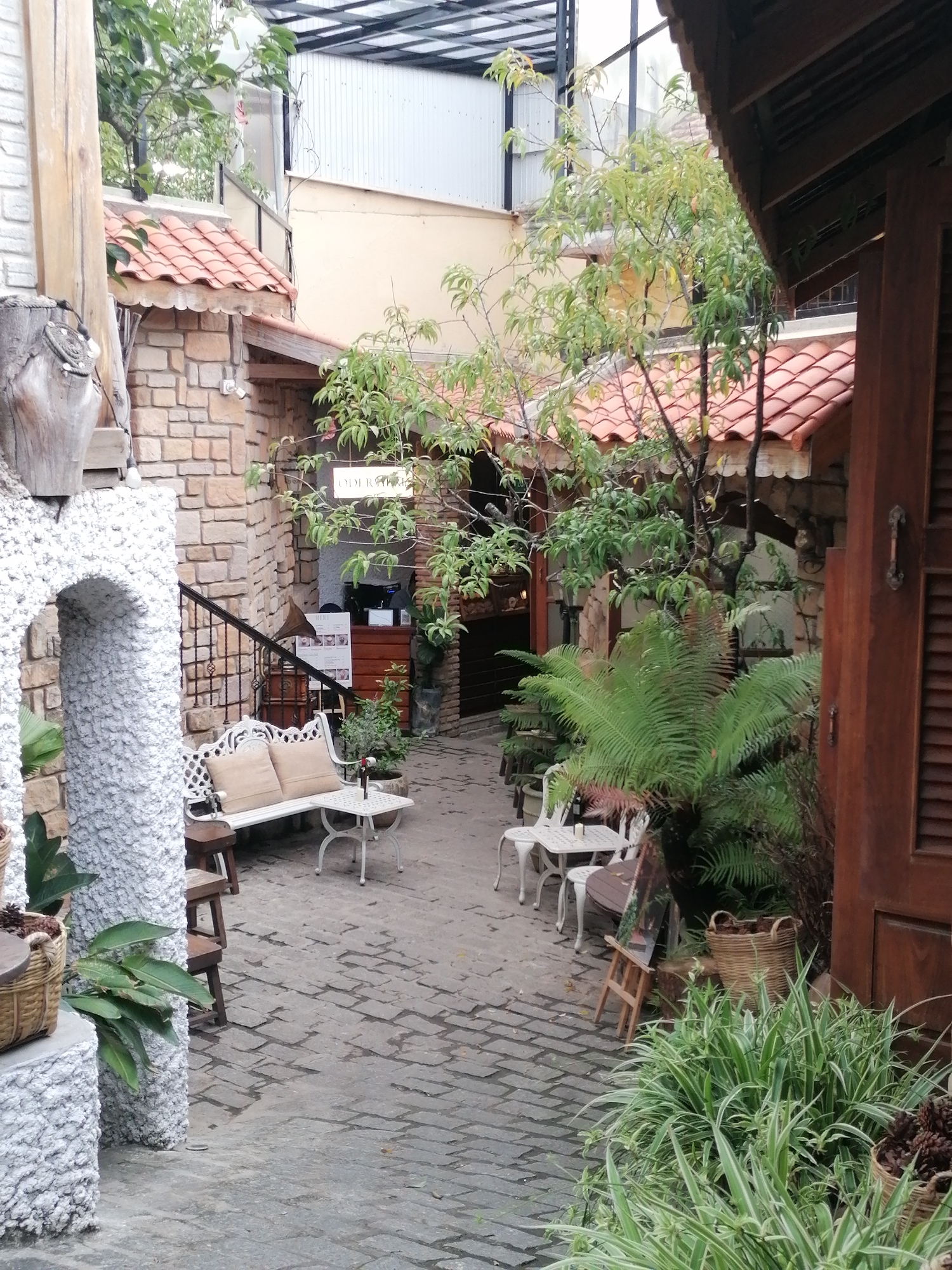 The image depicts a charming, secluded courtyard with a rustic and inviting ambiance. The scene features a cobblestone pathway leading into a covered outdoor seating area. To the left, a white stone archway frames a glimpse of lush greenery, with a large wooden beam and decorative pottery suggesting an artisanal or garden-themed establishment. The walls are constructed of light-colored brick, and the roof is tiled with terracotta.
Several pieces of white metal outdoor furniture are arranged in the courtyard, including a long bench with two cushions, a small table with chairs, and another table with two chairs. A white table with an umbrella stands near the bench. Plants are abundant, with ferns, leafy bushes, and potted greenery adding to the natural and tranquil atmosphere. A woven basket filled with pinecones is visible on the left side of the image.
In the background, a sign hangs from a tree branch, displaying white text on a black background that appears to read "ODERTHI". This, along with the architecture and lush vegetation, hints at a cafe or restaurant setting. The overall mood is peaceful and relaxing, suggesting a quiet afternoon in a pleasant climate. The lighting indicates it is daytime, likely with overcast skies or diffused sunlight, as there are no strong shadows. The presence of a wine bottle on one of the tables suggests leisurely dining or a social gathering. The environment evokes a sense of escape and tranquility, perhaps a hidden gem in a Vietnamese city like Da Lat, known for its cool climate and European-inspired architecture.