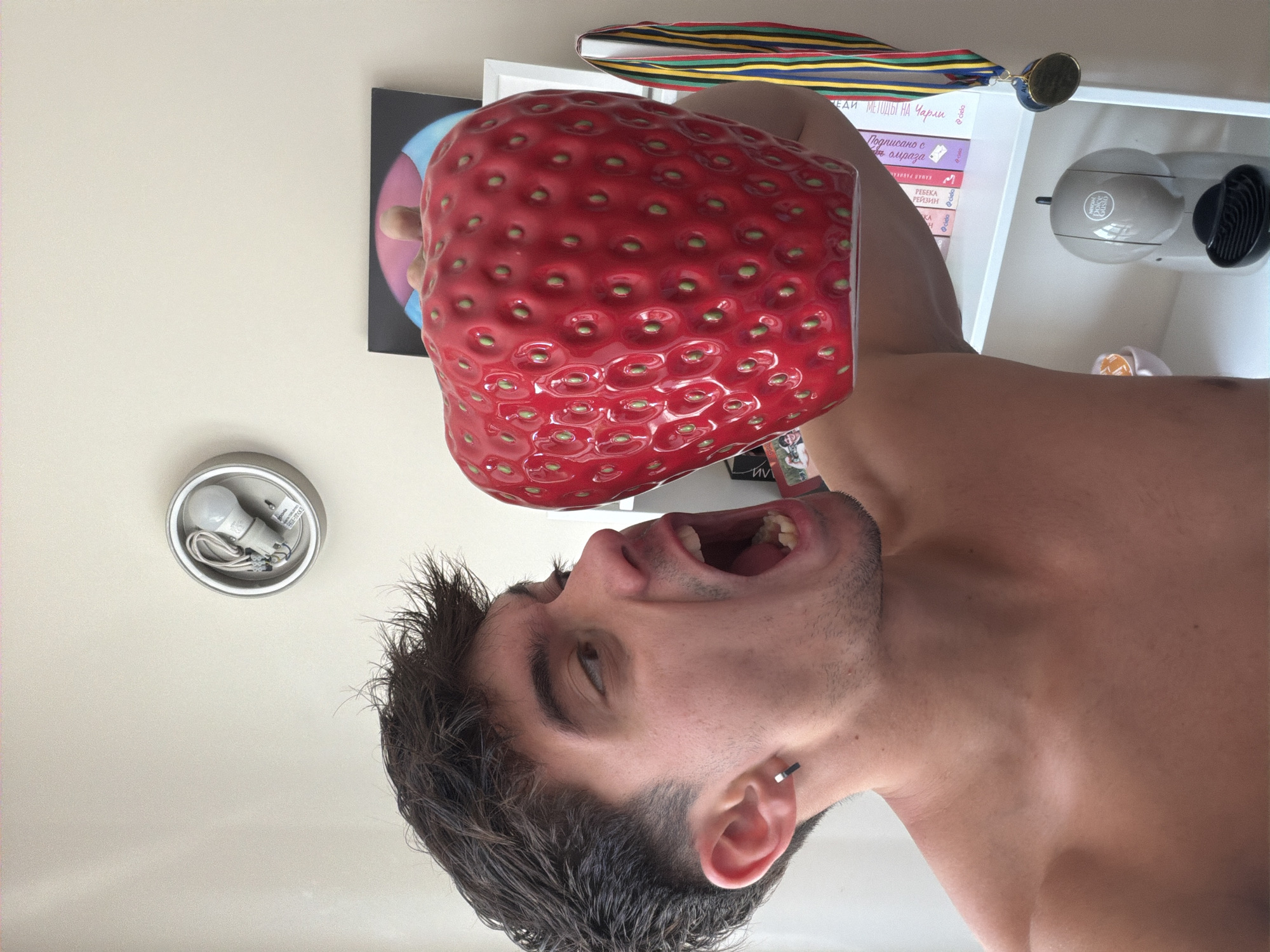 Here's a forensic description of the image:
**General Description:** The image shows a man appearing to attempt to eat a large strawberry-shaped object. The image is rotated counter-clockwise, suggesting it was taken with the camera held sideways or the phone was rotated after capture.
**People:** A young man is visible from the chest up. He has dark hair, dark eyebrows, and appears to have a small earring. His mouth is wide open, seemingly about to bite into the object.
**Objects:**
* A large, artificial or decorative strawberry is positioned directly above the man's mouth.
* Behind the strawberry, a white shelf with a coffee maker and several books.
* A medal hanging on the upper shelf.
* On the ceiling, a light fixture with a light bulb is visible.
* A framed picture is partially visible to the left of the shelf.
**Environment:**
* The setting appears to be inside a home or apartment, based on the wall color and the items in the background.
* The visible books contain Cyrillic characters indicating the location in which the picture was taken is likely Bulgaria. The books titles suggest the location of the image is in Sofia, Bulgaria.