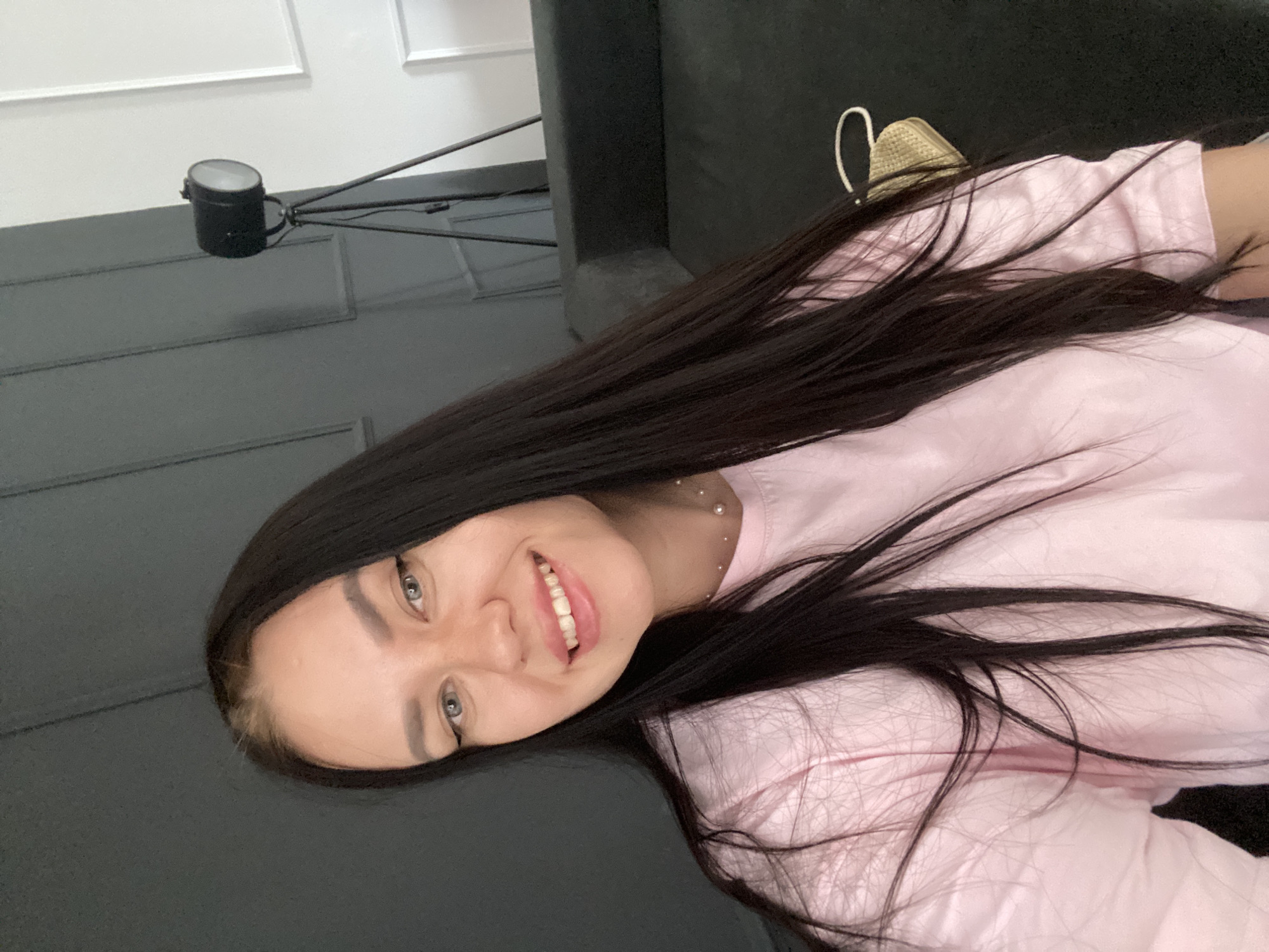 A young woman with long, dark hair is looking at the camera and smiling. She is wearing a pink t-shirt. In the background, there is a dark gray couch, a black spotlight on a tripod, and a wall with gray paneling and white molding. The image appears to have been taken indoors, possibly in a home or studio setting. There is no visible indication that this scene is located in the city of Chaikovsky, Russia.