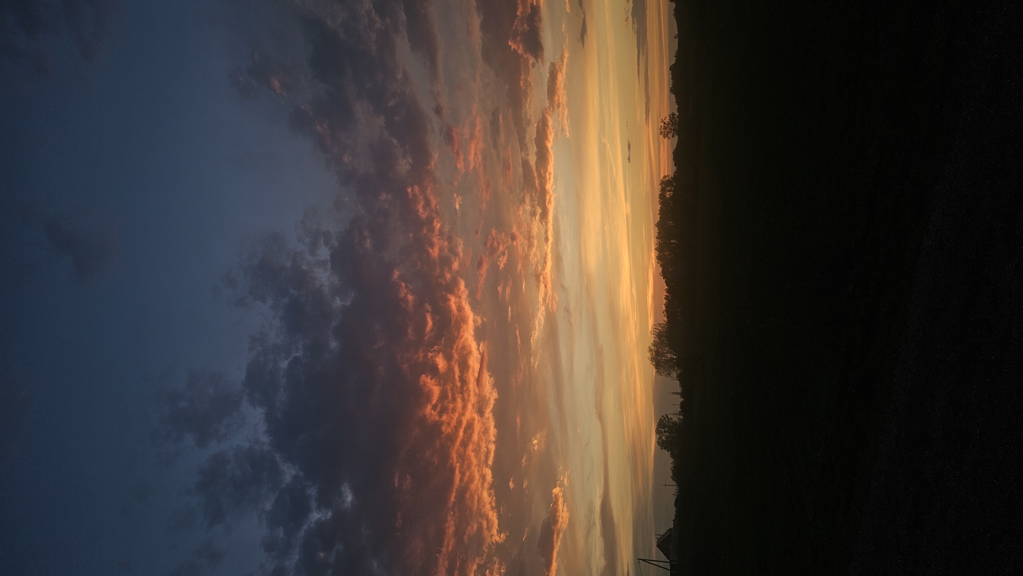 The image depicts a vivid sunset over Yangiskain, Russia. The sky is dominated by clouds with fiery orange and reddish hues, transitioning to shades of blue and grey. A dark silhouette of trees and land occupies the lower right portion of the frame, creating a stark contrast with the vibrant sky.