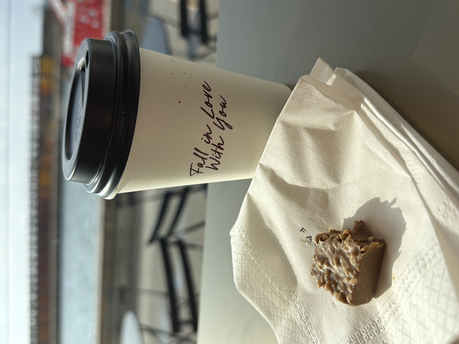 The image presents a close-up, slightly elevated view of an outdoor cafe table in Tel-Aviv, Israel, with a body of water and a distant structure in the background, consistent with the Tel Aviv Port (Namal Tel Aviv) area. In the foreground, on a dark grey table surface, a white disposable coffee cup with a black plastic lid is positioned to the right. The cup is inscribed with the dark brown text "Fall in Love With You". To the left of the cup, a folded white paper napkin with a subtle patterned border lies flat. Resting on the napkin is a rectangular piece of chocolate, appearing to contain nuts or crispies, with a visible bite mark on its right side, indicating partial consumption. Natural sunlight from the left casts distinct shadows from the cup and chocolate onto the table and napkin. The blurred background reveals an expanse of blue-grey water, beyond which a long, dark, linear structure, resembling a pier or breakwater, extends horizontally. Out-of-focus black outdoor cafe tables and chairs are also faintly visible behind the primary subjects. No individuals are present in the scene.
