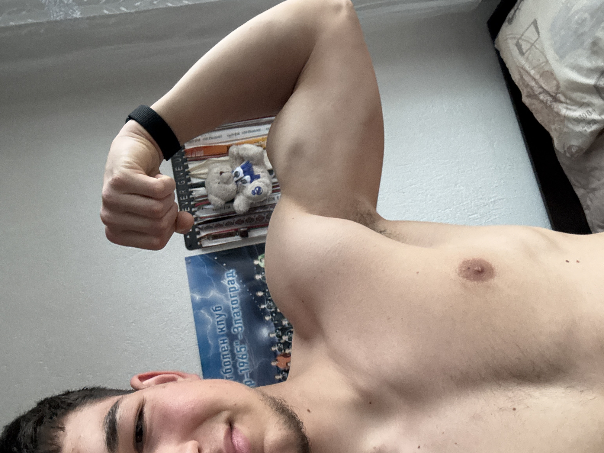The image captures a young male posing indoors, seen from a low, upward-looking angle. He is shown from the chest up, with his face partially visible in the bottom left. He has short dark hair, some facial stubble, and a neutral to slightly smiling expression. His left arm is raised and flexed, showcasing his bicep, and he wears a black wristband. His torso is bare, revealing his chest and shoulder.
In the background, behind his flexed arm, two small, light-colored teddy bears are stacked on top of what appears to be books or DVDs. One of the bears has a blue accessory. Partially visible text on these items reads "EVER GIVE U", likely part of "NEVER GIVE UP".
A blue poster is affixed to the wall behind the person's head and torso. Visible text on the poster includes "футболен клуб" (football club) and "1965 – Златоград", indicating the location is Zlatograd, Bulgaria. In the upper right corner, a section of a bed with patterned bedding is visible. The scene is illuminated by soft, even indoor lighting.