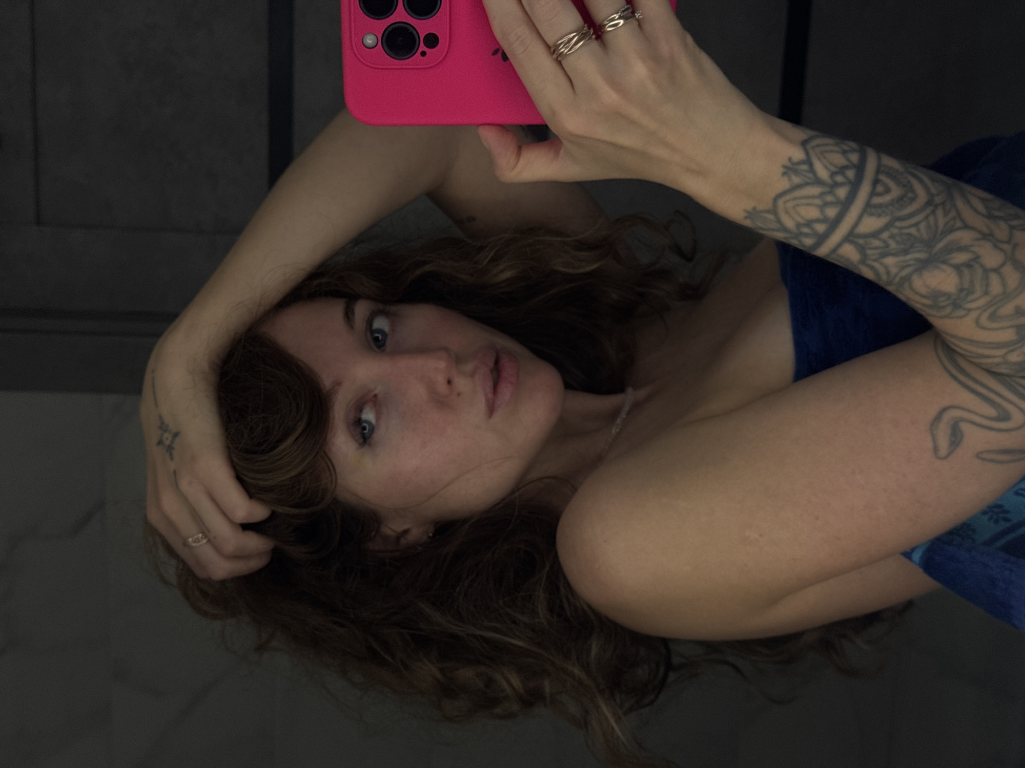 A female individual is depicted in a close-up, waist-up portrait, appearing to take a selfie reflection in a mirror. She has long, wavy brown hair, blue eyes, and fair skin. A small yellowish discoloration is visible beneath her left eye. Her left arm is raised, with her hand positioned near the top of her head, fingers interlocked with her hair. A simple ring is on her left ring finger, and a small star-shaped tattoo is on the back of her left hand. She is wearing a dark blue towel wrapped around her torso. Her right arm extends forward, holding a bright pink smartphone with a visible triple-lens camera array and a partially obscured Apple logo, consistent with an iPhone Pro Max model. Multiple gold-toned rings are on her right ring and middle fingers. Her right forearm is extensively tattooed with an intricate mandala-style sleeve design, including a serpentine element on the outer bicep area. A thin, light-colored beaded necklace is visible around her neck. Her gaze is directed slightly off-camera to her left, with her lips slightly parted. The background is a dark, indistinct indoor setting, possibly a wall or tiled surface, with minimal discernible features.