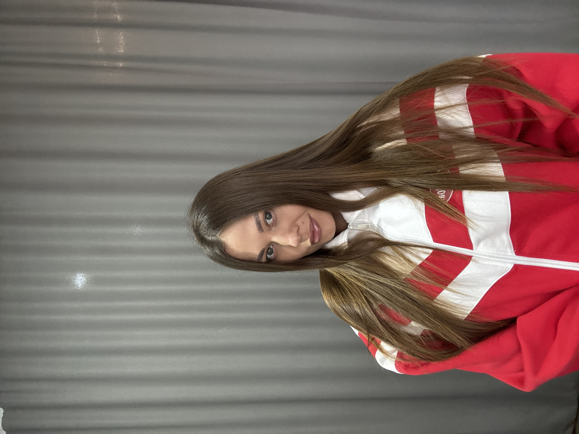 A brunette woman wearing a red and white jacket is positioned diagonally in the frame. The background is a grey, horizontally textured wall. The location is Chernushka, Russia.