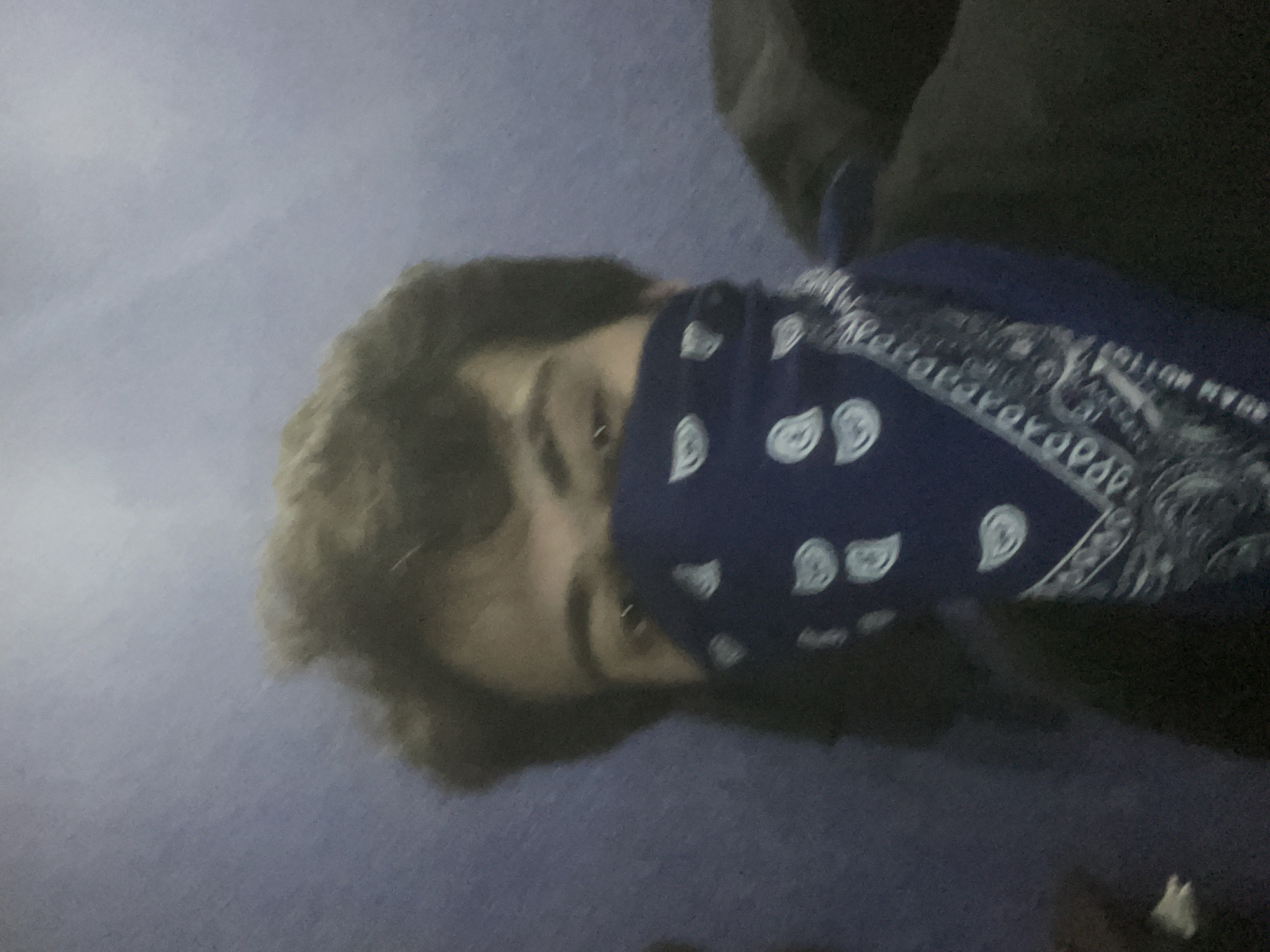 In the foreground of the image, a person's face is partially obscured by a dark blue bandana with a white paisley pattern. The bandana covers the lower half of their face. Visible above the bandana are the person's eyes, eyebrows, and brown hair. The background appears to be a solid blue surface, possibly a wall or fabric, with some slight texture. The lighting is dim, creating soft shadows and a somewhat grainy texture throughout the image. The image quality is low, with a lack of sharp focus. No specific location in the city of Tbilisi, Georgia, can be identified from the provided visual information.
