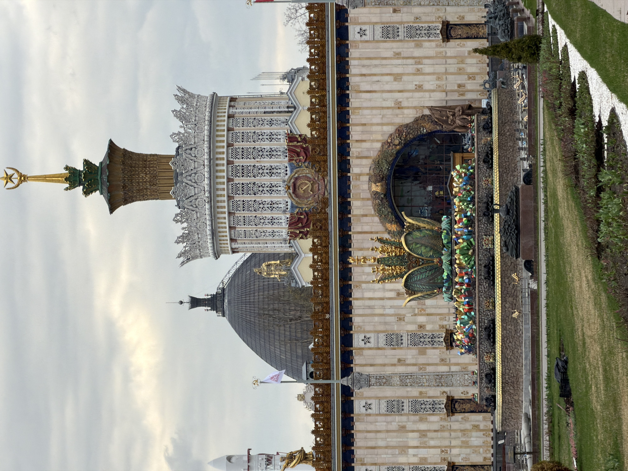 The image displays a grand architectural complex, likely a pavilion within a park or exhibition grounds, viewed under an overcast sky. The dominant structure on the right features a tall, golden spire culminating in a crescent moon and star finial. This spire rises from an ornate, multi-tiered cylindrical base adorned with intricate white and gold lacework patterns.
Below this, the building's facade is constructed of light-colored, patterned blocks, interspersed with decorative elements and golden railings. A prominent feature is a large arched entrance or window, framed by vibrant, colorful, mosaic-like decorations. Centered above this arch is a distinct Soviet-era emblem: a golden hammer and sickle within a wreath, set against a red background. The base of the building below the arch is further embellished with a variety of colorful, abstract decorative shapes and textures.
To the left and slightly behind this main section, a large, dark-grey domed building is visible, featuring golden statues on its upper reaches. A white flag, bearing a red logo with discernible text "ВДНХ" (VDNKh) and "МОСКВА" (Moscow), flies from a flagpole positioned near this dome.
The foreground depicts a meticulously maintained lawn, interspersed with rows of hedges or small trees, and delineated by white pathway borders. The sky is uniformly overcast, indicating daytime with diffused light. No people or specific activities are present, leaving the focus entirely on the monumental architecture and the serene, landscaped environment. The visible Soviet emblem, alongside the opulent architectural style, strongly suggests the location is VDNKh in Moscow, Russia.