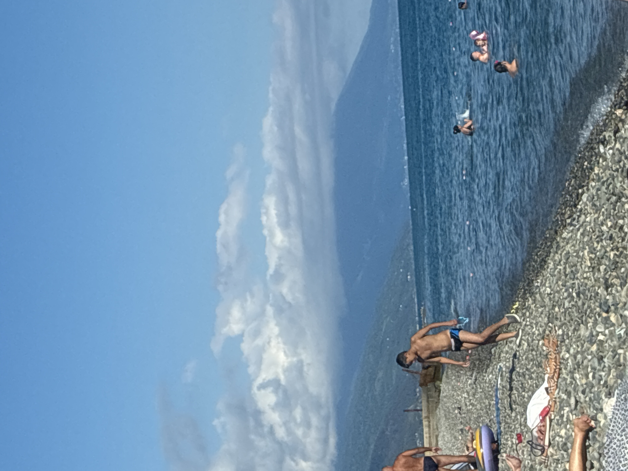 The image depicts a sunny day at a pebble beach in Sirius, Russia, adjacent to the Black Sea. In the foreground, the pebbled shore is occupied by several people. A young boy in blue swim trunks is walking towards the water, holding a pair of goggles. To his left, a man in black swim trunks stands facing away from the camera. Further left, a person is partially visible lying on a yellow and purple inflatable float. Beach towels and personal belongings are scattered on the pebbles. The sea, a deep blue, is populated with swimmers. Several individuals are seen enjoying the water, some with inflatable toys. In the background, a chain of green mountains rises, with some cloud cover obscuring the peaks, under a bright blue sky dotted with white clouds.