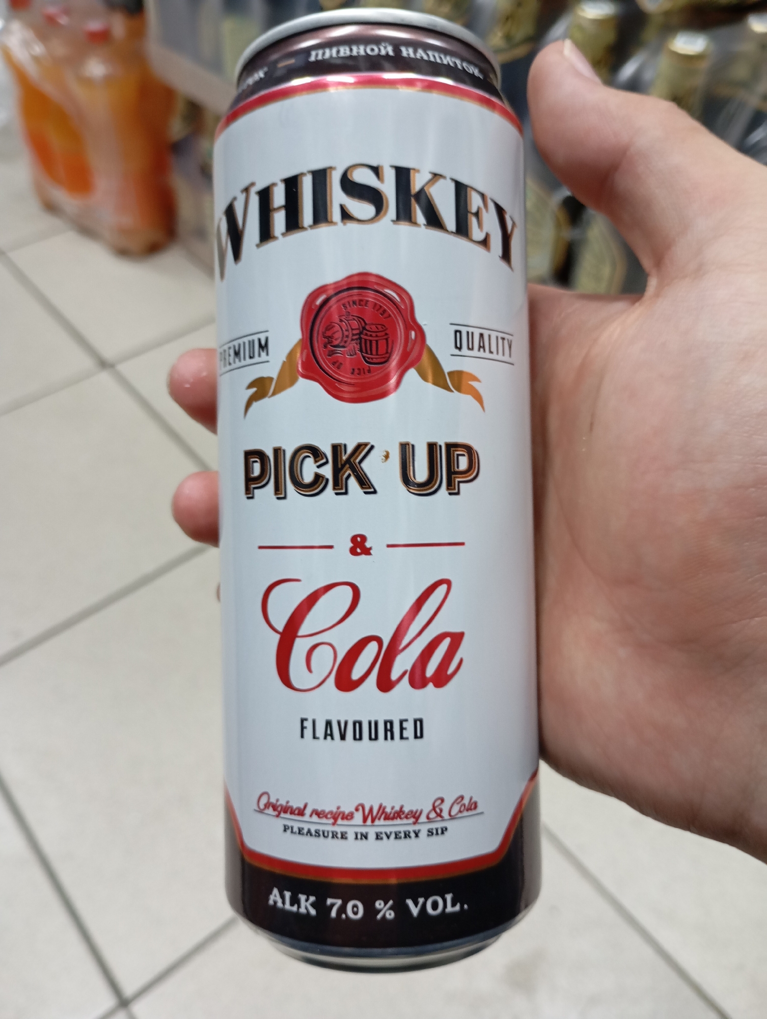 A person's hand holds a can of "WHISKEY PICK UP & Cola" beverage. The can is white with black and red lettering and a red seal. The background appears to be a store aisle with shelves of other beverages, suggesting the can was recently purchased or is being viewed in a retail setting. The flooring appears to be tiled. The scene is indoors, in Moscow, Russia.