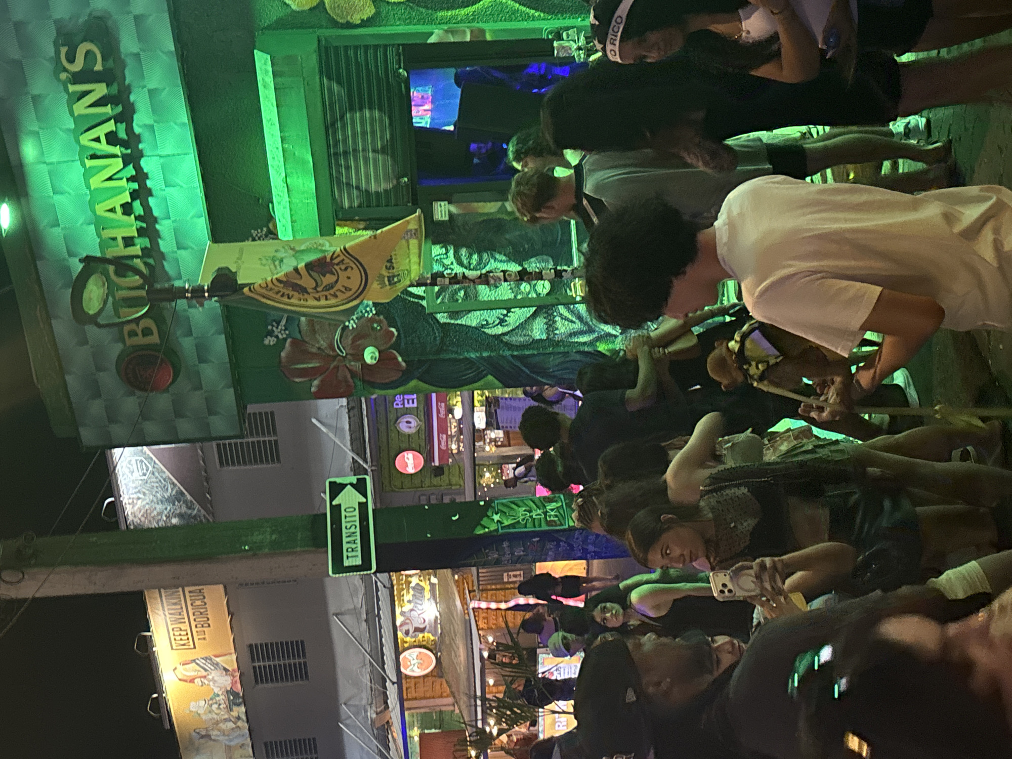 The image depicts a nighttime street scene in San Juan, Puerto Rico. A crowd of people is gathered outdoors, with several businesses lining the street. In the foreground, a woman is holding a smartphone to take a picture. She is wearing a black top and a dark skirt. To her right, another woman is visible, wearing a white top and dark shorts, with a headband that says "PUERTO RICO." Behind them, various individuals are seen in casual attire.
The environment is illuminated by bright green lights, primarily from a sign that reads "BUCHANAN'S." Other businesses with signage are visible, including one with a red Coca-Cola logo and another that appears to be a bar or restaurant with an awning. A green traffic sign with an arrow and the word "TRANSITO" is mounted on a pole. The overall atmosphere suggests a lively urban nightlife setting.