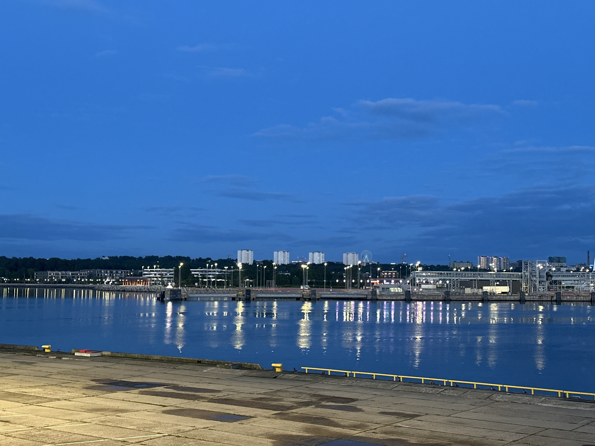 The image depicts a waterfront scene in Tallinn, Estonia, likely at dusk or early morning, given the twilight sky and artificial lights. The foreground features a concrete pier or dock with some patches of moisture reflecting the ambient light. A yellow railing runs along the edge of the pier.
Beyond the pier, a calm body of water, presumably the Baltic Sea, stretches towards the city skyline. The water reflects the numerous lights from buildings and streetlights lining the shore. These reflections create a shimmering effect across the water's surface.
In the mid-ground, a series of buildings and infrastructure line the waterfront. These include modern apartment buildings, commercial structures, and what appears to be port facilities with cranes and walkways. A Ferris wheel is visible in the distance, suggesting an entertainment area or landmark within the city. The sky above is a deep blue, with scattered, wispy clouds.
The overall impression is one of a tranquil, urban waterfront at the transition between day and night. No people are visible in the image.