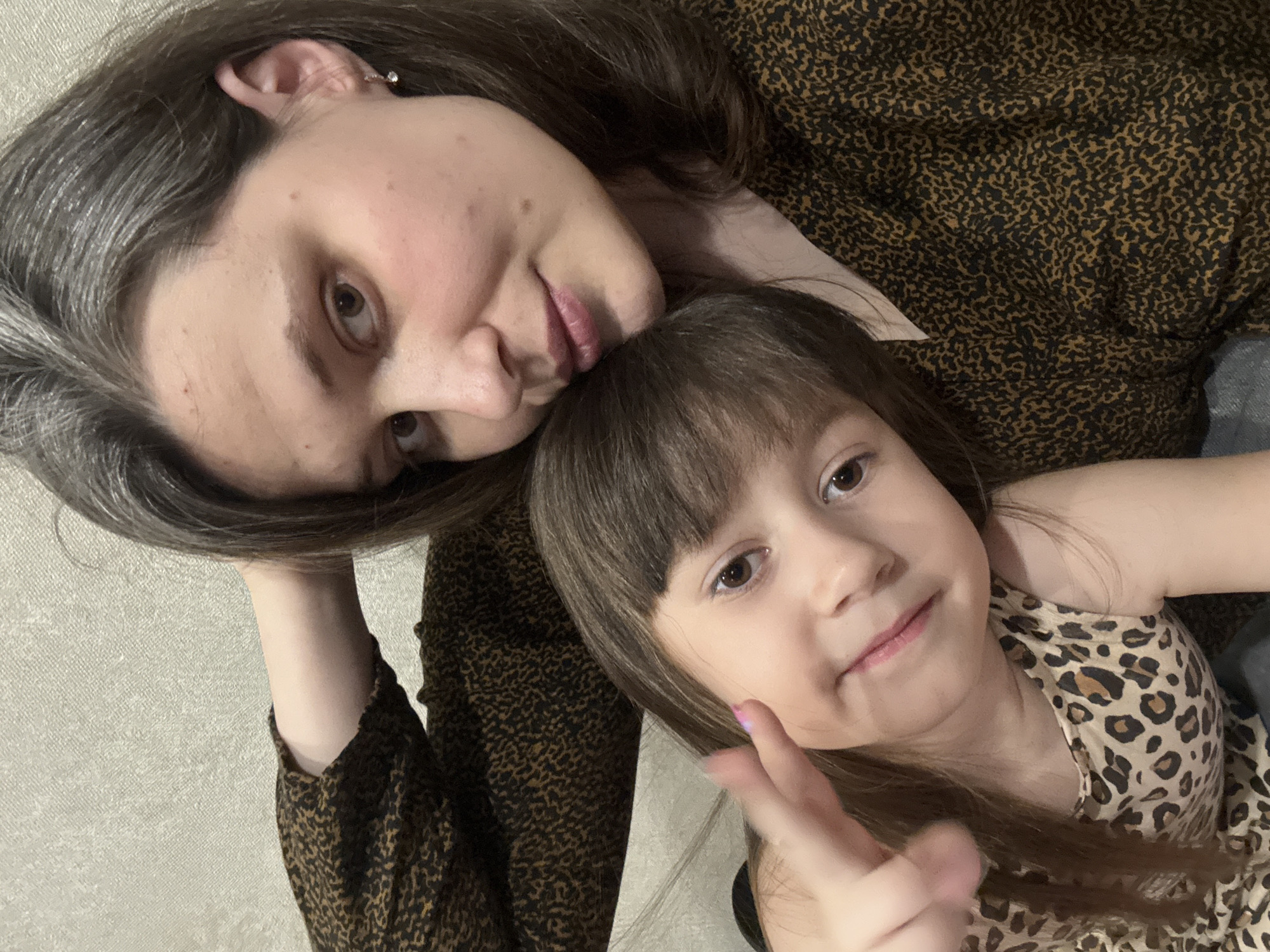 A woman and a child are posing for a selfie. They are lying down, close to each other. The woman is wearing a brown animal-print shirt, and the child is wearing an animal-print tank top. The child is holding up a peace sign with their fingers. The location may be Chaikovsky, Russia, but there is no visible evidence within the image to confirm this location.