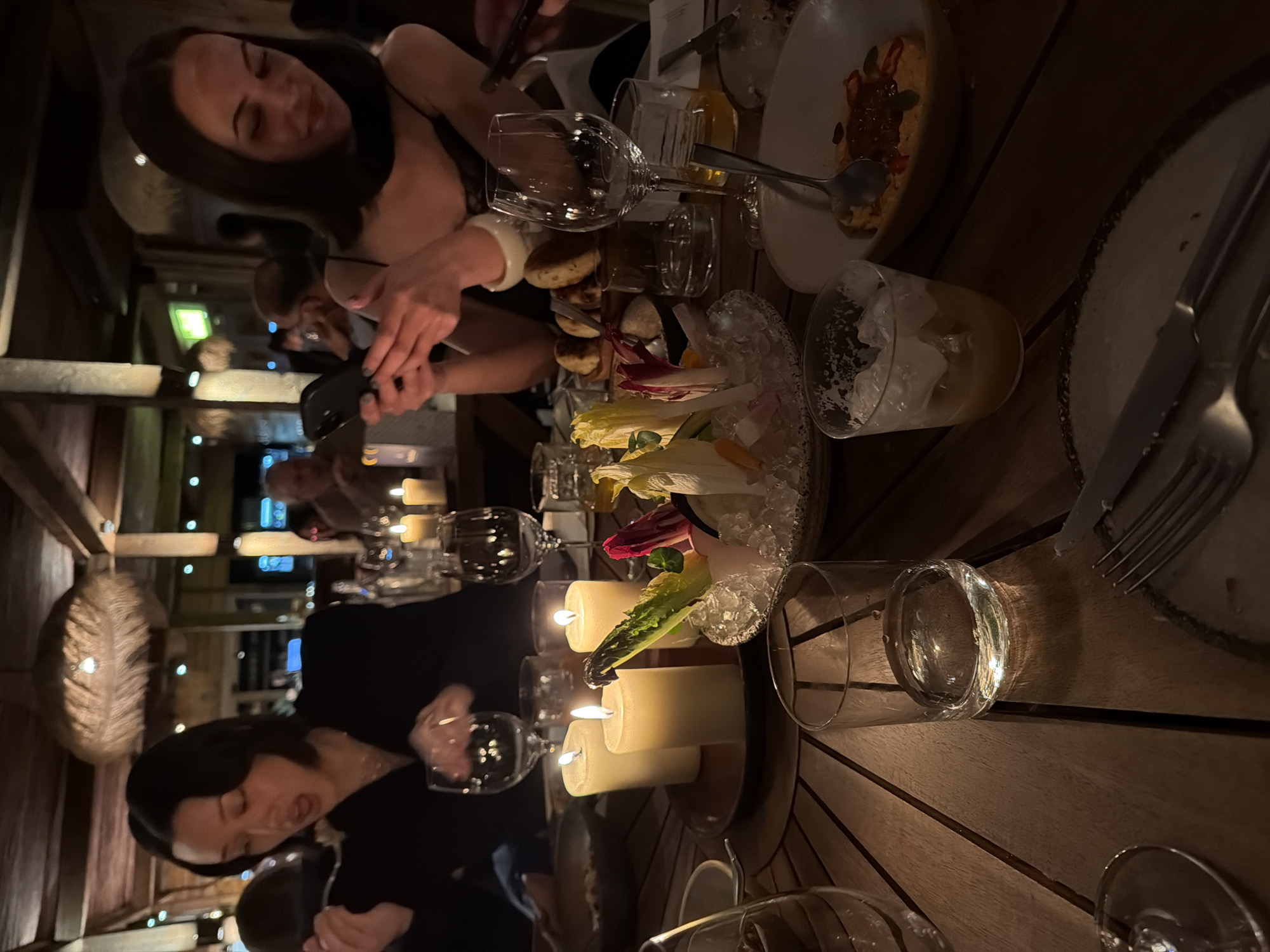 The image depicts a group dining in a dimly lit restaurant with wooden tables, likely located in London, United Kingdom. At least two women are visible in the frame, with one actively taking a photo with her phone. The table is set with several courses, including a salad served on ice, appetizers, and partially consumed plated dishes. Various glassware items like wine glasses and water glasses are scattered across the table, alongside cutlery and lit candles. The environment suggests a contemporary setting with a focus on dining ambiance, as evidenced by the dim lighting and decorative elements. The overall atmosphere seems relaxed and informal, possibly a social gathering among friends.