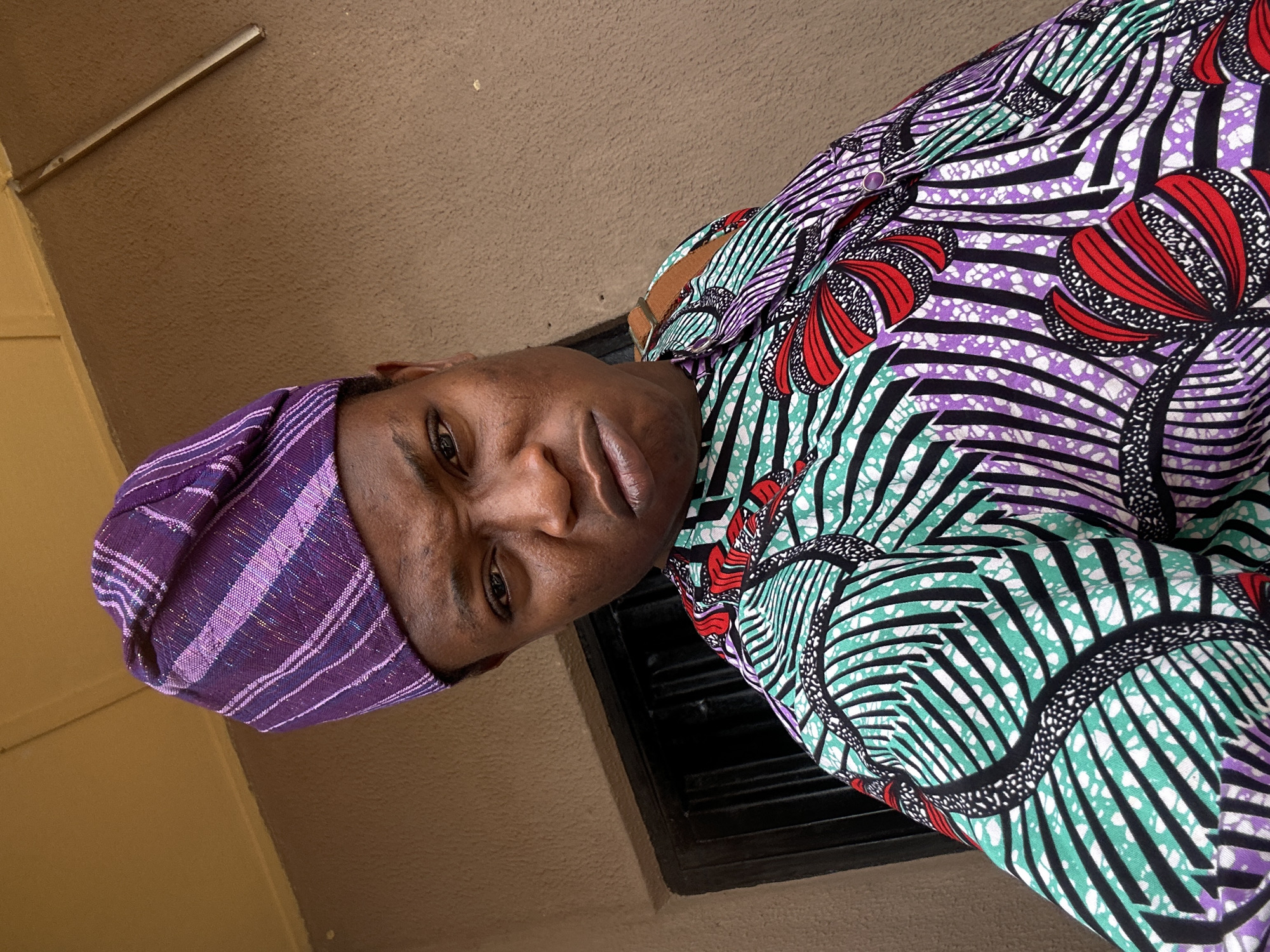 A medium, tilted, eye-level shot depicts a person wearing a purple headwrap and a colorful, patterned shirt with shades of purple, red, green, black, and white. The background includes a wall and a dark, square-shaped object, possibly a window. The setting appears to be indoors, but the exact location within the city of Unknown, Unknown cannot be determined from the image.