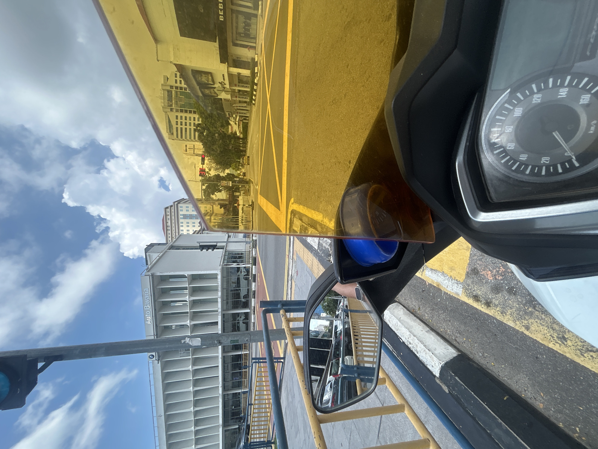 This is a first-person perspective, eye-level, medium shot image of a motorcycle rider at an intersection in George Town, Malaysia, on a bright, partly cloudy day. The rider is stopped, indicated by the presence of a traffic light showing red. The foreground is dominated by the motorcycle's dashboard with a speedometer indicating approximately 10 km/h and part of the handlebars. A yellow tinted windshield partially obscures the view ahead.
The background shows a modern, white building with a repeating grid facade, labeled "Auto Bavaria," a car dealership. To the right, another building displays "BEBE," suggesting a retail establishment. The street is paved with asphalt and marked with yellow lines. A yellow barrier and blue railings line the sidewalk. The motorcycle's side mirror reflects a line of parked cars. The overall scene depicts a typical urban streetscape.