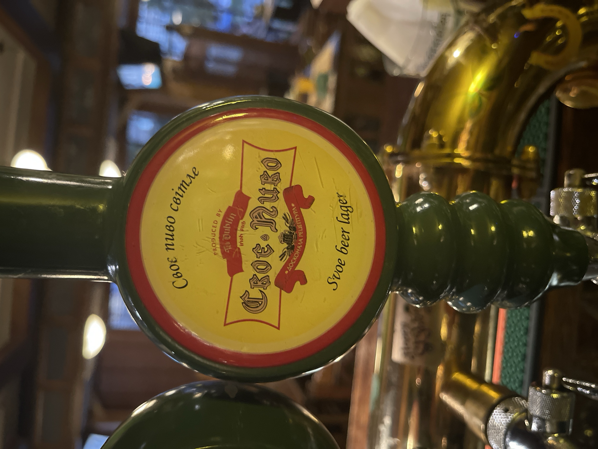 The image shows a close-up of a beer tap handle, likely in a pub or bar. The handle is green and features a circular emblem with a yellow background and red trim. The emblem displays text in what appears to be a Cyrillic-based alphabet, along with the words "Irish pub" and "Dublin" in English. Below this, there is more text in the Cyrillic-based alphabet and "Svoe beer lager." In the background, out of focus, there are tables, chairs, and lighting fixtures, suggesting an indoor establishment. The overall scene suggests a setting where beer is served.