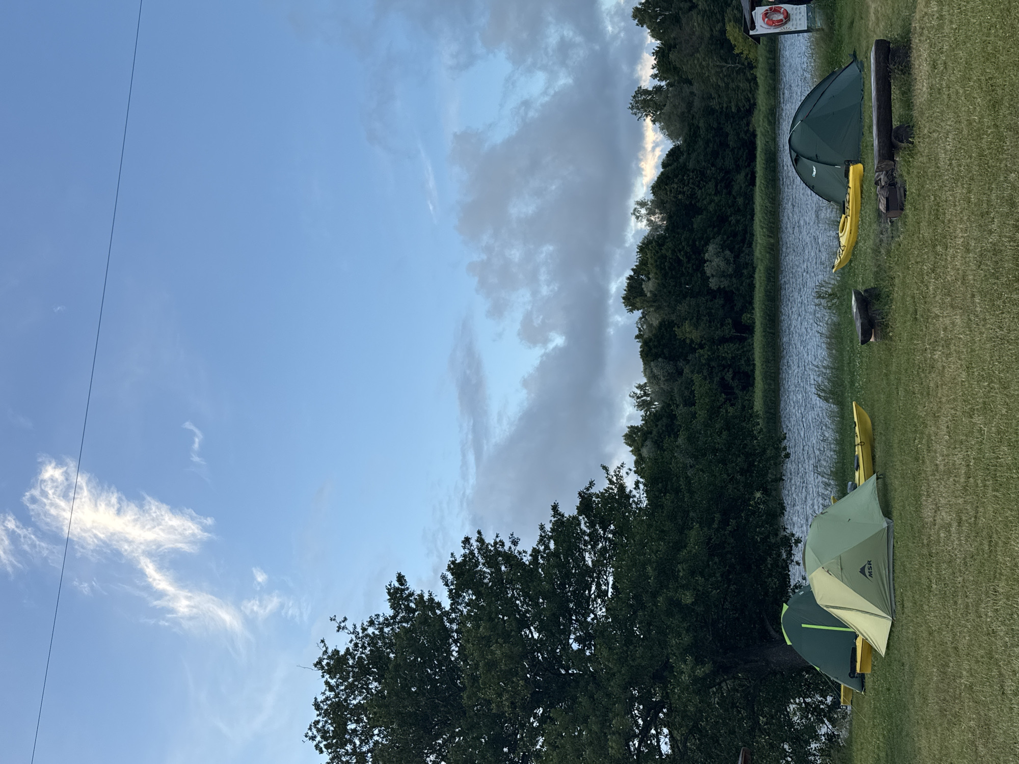 The image depicts a campsite by a body of water, likely a river or lake, in Carnikava, Latvia. In the foreground, a grassy area is occupied by three tents: two light green tents in the left foreground and a dark green tent on the right. Yellow kayaks are placed beside each tent. Two wooden logs serve as benches or seating, and a small campfire pit is visible between them. A large, mature tree with dense foliage stands to the left of the frame, casting a shadow over some of the tents. Behind the campsite, the water stretches to a treeline on the opposite bank, composed of various types of trees and vegetation. The sky above is partly cloudy, with streaks of white clouds against a blue background. A power line stretches diagonally across the upper portion of the image.