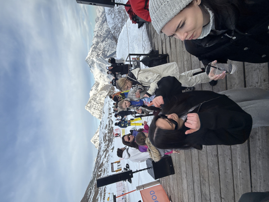 The image depicts an outdoor social gathering on a wooden deck in a snow-covered alpine setting, identified as Alagna Valsesia, Italy. The bright daytime scene is dominated by majestic snow-capped mountains under a partly cloudy sky.
In the foreground, two young women are prominent. On the right, a woman in a grey beanie and black winter jacket holds a clear plastic cup with ice, looking down introspectively. To her left, another woman with long dark hair, wearing sunglasses and a black jacket, is partially visible, making a hand gesture with both thumbs pointing downwards. Behind them, several people, mostly young adults dressed in ski wear and various winter clothing, are socializing, some seated and others standing.
A large black speaker on a stand is positioned on the wooden deck in the mid-ground, with another speaker partially visible further back. The deck itself is constructed from wooden planks. The setting is a high-altitude terrace or viewing platform amidst a vast, rugged mountain range, typical of a ski resort.
The activity appears to be an apres-ski event or an outdoor party/concert, characterized by the presence of music equipment and people enjoying drinks and conversation in the chilly environment. The weather is cold and clear, with abundant snow covering the ground and peaks. The time of day is daylight. The woman in the beanie shows a pensive expression, while the other woman appears more animated. The location cues strongly suggest a winter sports destination.
The word "WOOD" is partially visible in white lettering on an orange sign or box near one of the speakers on the deck.