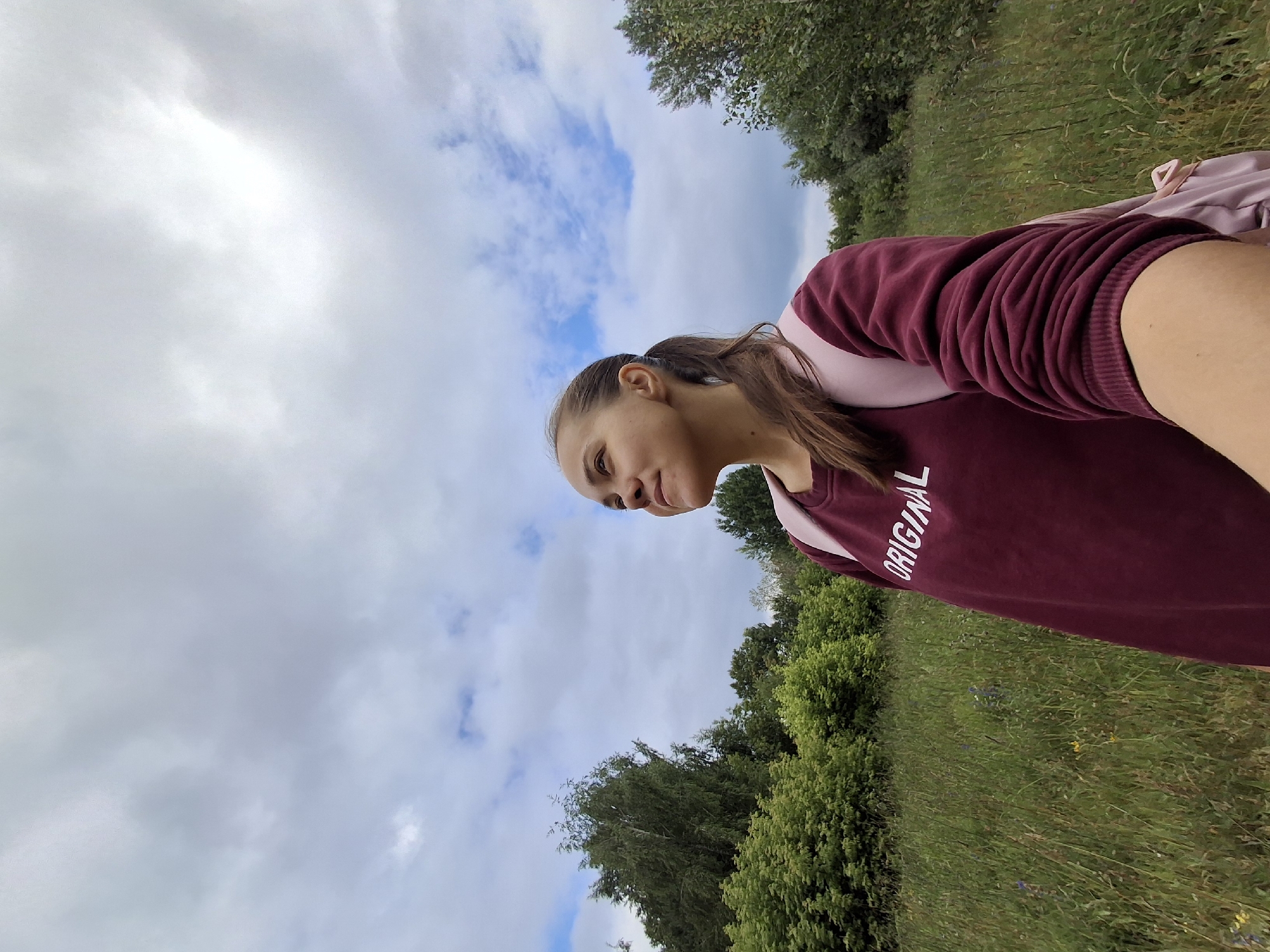 A young woman with her hair in a ponytail, wearing a maroon sweatshirt with the word "ORIGINAL" printed on it and a pink backpack, stands in a grassy field under a cloudy sky. She is looking upwards and to her right. The background consists of a dense line of trees and bushes, with a few scattered small plants in the foreground. The location is identified as Chaikovsky, Russia.