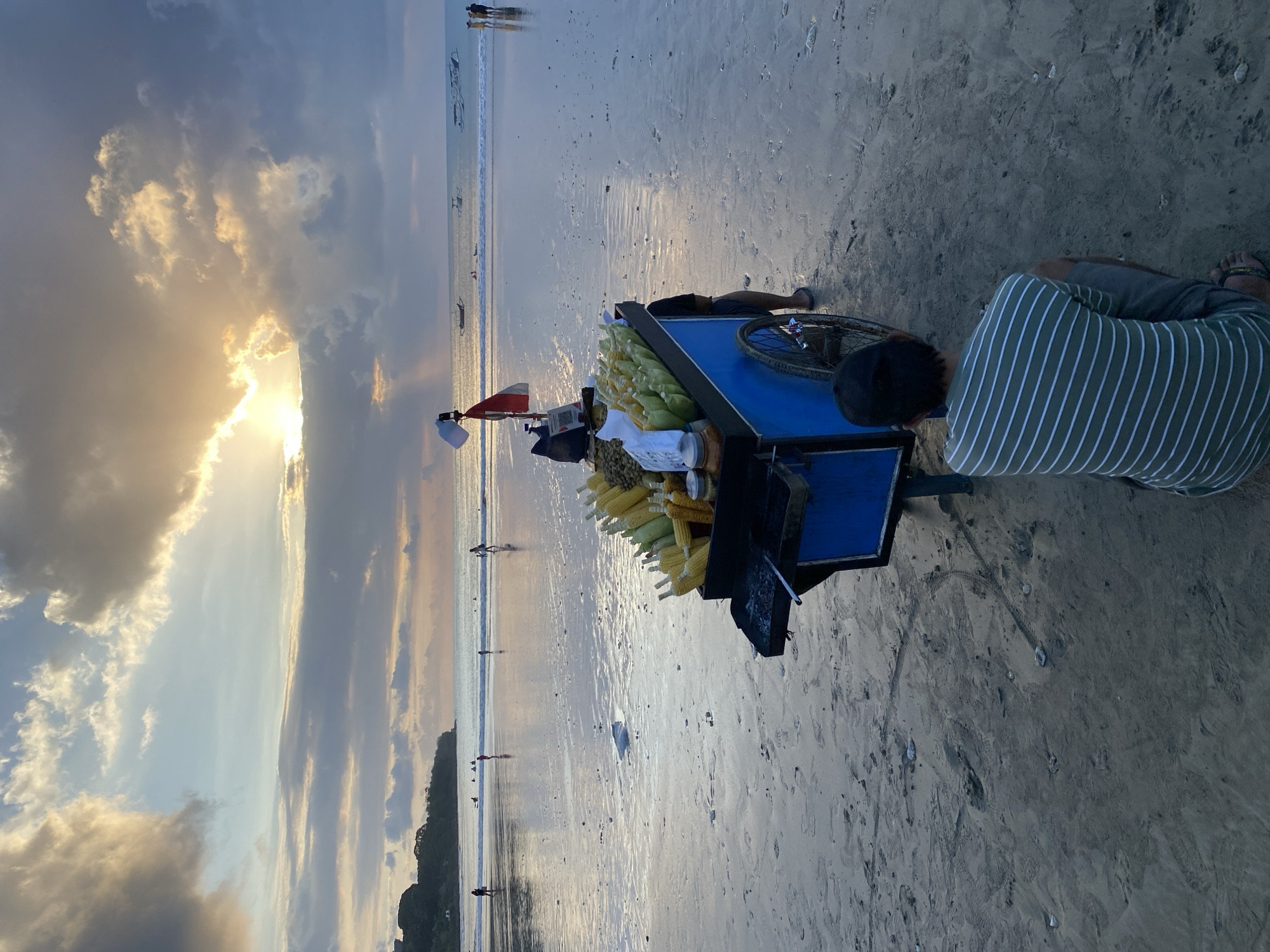 The image, oriented on its side, depicts a serene beach scene at either sunset or sunrise, illuminated by a dramatic golden glow emanating from behind scattered clouds. The setting is identified as Kuta Selatan, Indonesia.
In the foreground, a vendor is partially visible, seen from above and behind, kneeling or crouching on the wet sand. They are wearing a green and white striped shirt and dark shorts. Next to them is a blue vendor cart, equipped with a wheel, laden with numerous yellow corn on the cob, some still wrapped in husks, and what appear to be bottled drinks or packaged items. A small red and white flag is attached to the cart, possibly an Indonesian flag.
The sandy beach, wet from the receding tide, reflects the warm sky, and gentle waves are seen breaking along the shore. Further out in the water, several small, dark boats, possibly traditional vessels, are visible. In the mid-ground and distance, numerous small figures of people are scattered along the waterline, walking or wading, enjoying the coastal atmosphere. The sky features a mix of soft blues, greys, and striking golden light where the sun breaks through the clouds. Distant land or hills can be faintly seen on the horizon. No discernible text is visible within the image. The overall impression is one of calm and leisure typical of a beach evening or morning.