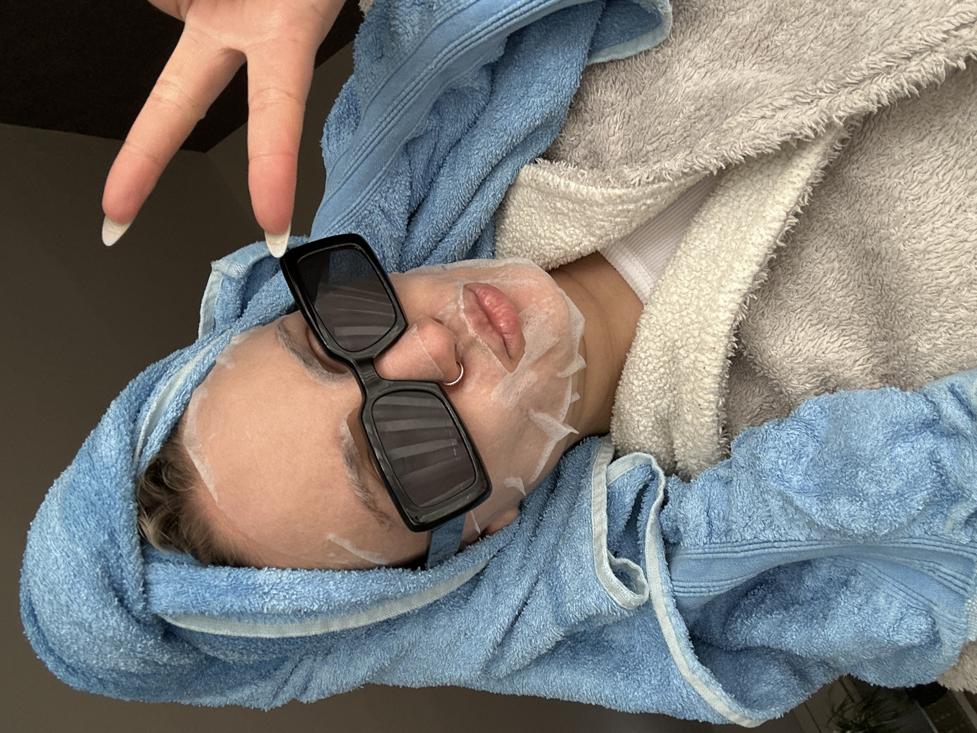 The image presents a rotated close-up of a person engaged in a self-care routine. The individual's head is oriented towards the left of the frame, wrapped in a light blue terry cloth towel. A white sheet face mask covers most of their face, with cutouts revealing their lips and nostrils. Black, rectangular sunglasses are perched on the bridge of their nose, adding a playful touch. A small silver hoop nose ring is visible on their left nostril.
Part of a fluffy, light grey or off-white towel or robe is draped around their body. In the upper part of the frame, a hand with long, white-tipped manicured nails is making a peace sign. The background is dark and out of focus, suggesting an indoor setting. The overall impression is one of relaxation and self-indulgence. There is no visible text in the image, and no specific location cues for Esenyurt, Turkey, are discernible.