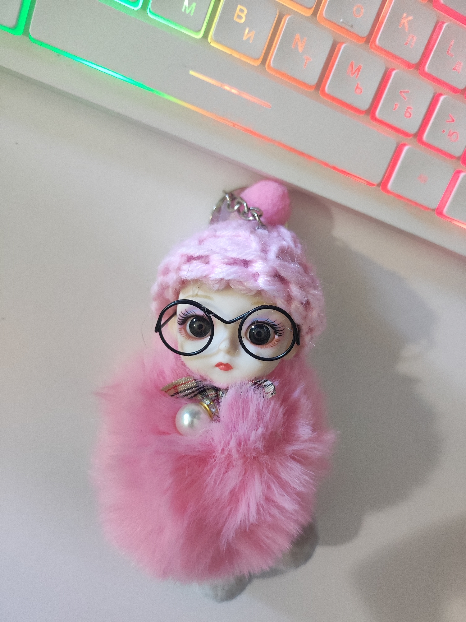 The image shows a close-up view of a keychain in the shape of a doll, which is placed on a white surface. The doll is wearing a pink knitted hat, large round glasses, and a fluffy pink body. It has a small pearl accessory attached to its chest. In the background, a portion of a keyboard with illuminated keys is visible. The keyboard displays a gradient of colors, from green and yellow to pink, suggesting a gaming or customized setup. The lighting in the scene is soft, casting a gentle shadow on the right side of the image. The scene does not provide any information or context that would allow for its location to be determined as Cherkasy, Ukraine.