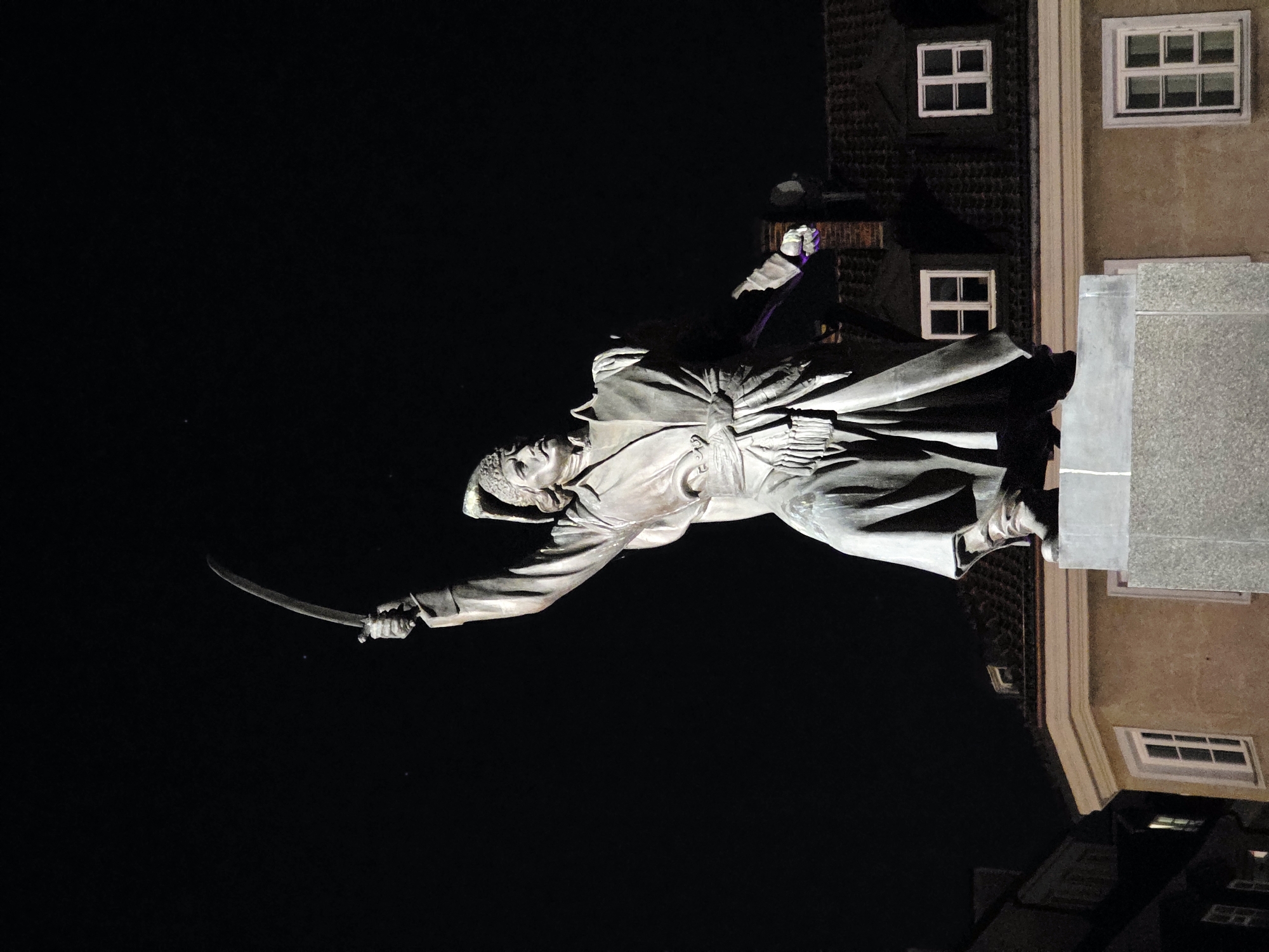 This is a nighttime, vertical-orientation photograph of a statue of a man in historical clothing, possibly a soldier or warrior, holding a curved sword. The statue is made of a bronze-like material and is illuminated by artificial light, creating strong shadows. The man is depicted in a dynamic pose, with his left arm extended forward, holding the sword in a sweeping motion. He wears a hat and a long, flowing coat.
Behind the statue, a building with multiple windows is visible. The building has a brick-like texture on its upper portion and a smoother, tan facade on the lower part. The windows are rectangular and appear to have multiple panes. The overall setting suggests an urban environment, possibly a public square or monument. The time of day is clearly night due to the dark sky and artificial lighting. There are no discernible weather conditions or strong emotional cues conveyed by the statue itself, other than the implied action. No text is visible in the image. The context provided indicates the location is Warsaw, Poland.