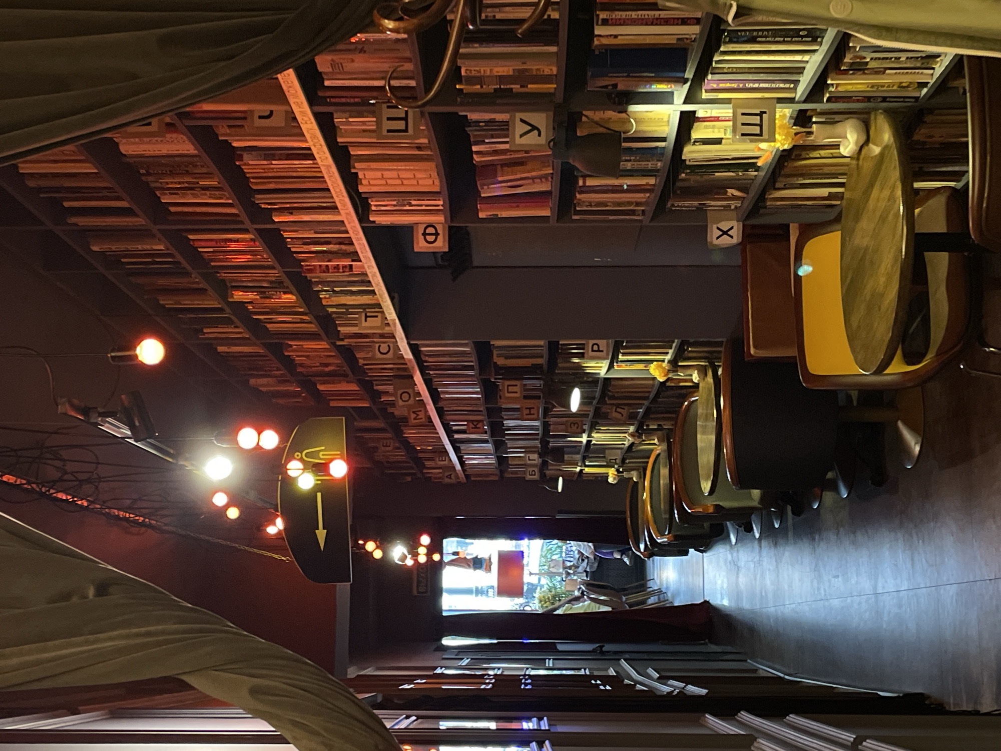 The image shows a corridor in a cafe/restaurant in Saint Petersburg, Russia. The walls are dark and lined with bookshelves filled with books, with Greek letters marking different sections. An arrow-shaped sign with a downward-pointing arrow and a symbol of a person using a restroom indicates the location of the restrooms. Chairs with yellow seating are lined up along one side of the corridor. String lights hang from the ceiling, illuminating the space. The end of the corridor leads to an entrance area with natural light and a view to the outside. A woman is visible near the entrance. A portion of a curtain or fabric is visible in the lower left corner.